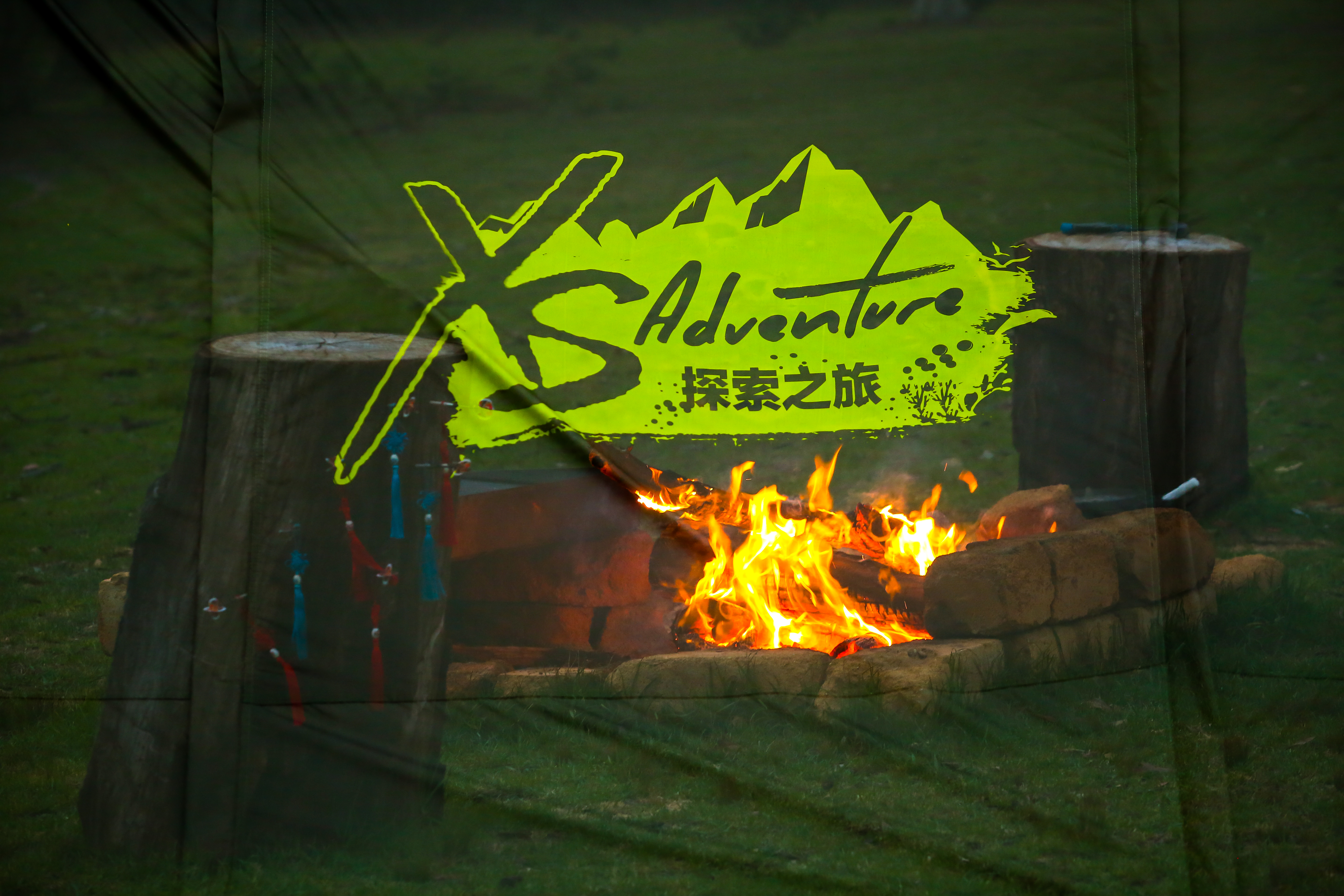 画面透过印有“XS Adventure 探索之旅”字样的活动标识幕布，可见户外草地上篝火正热烈燃烧，火焰明亮温暖，篝火旁摆放着原木树桩座椅，整体氛围自在鲜活，充分展现出XS探索之旅充满活力、亲近自然的活动特质，尽显轻松愉悦的户外拓展氛围。