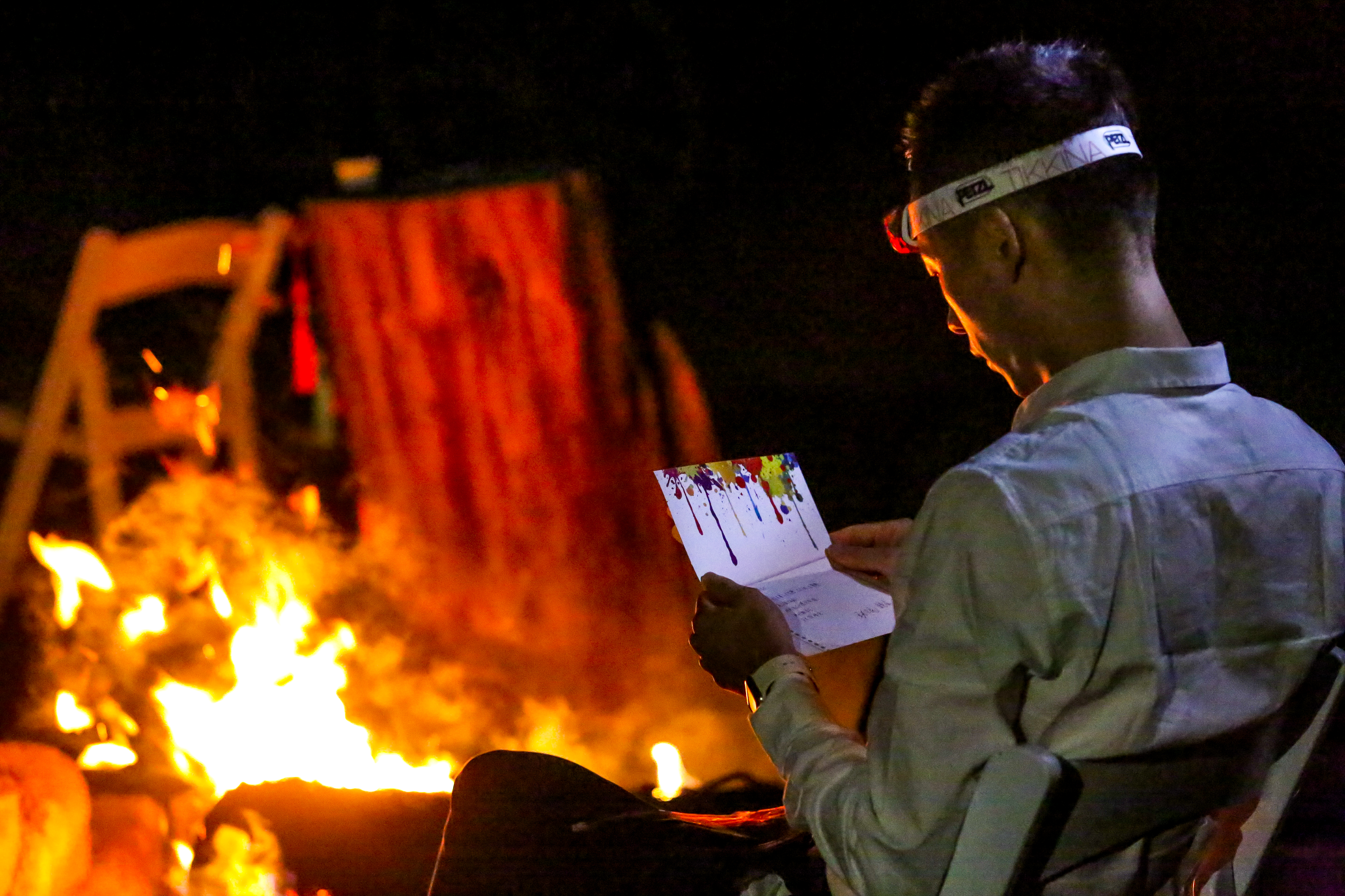 安利户外团建篝火活动现场，暖黄色篝火在夜色中跃动，一位佩戴头灯的伙伴正低头认真阅读带有泼墨装饰的活动定制手卡，氛围温馨治愈，尽显本次活动传递温暖、凝聚伙伴力量的核心初衷。