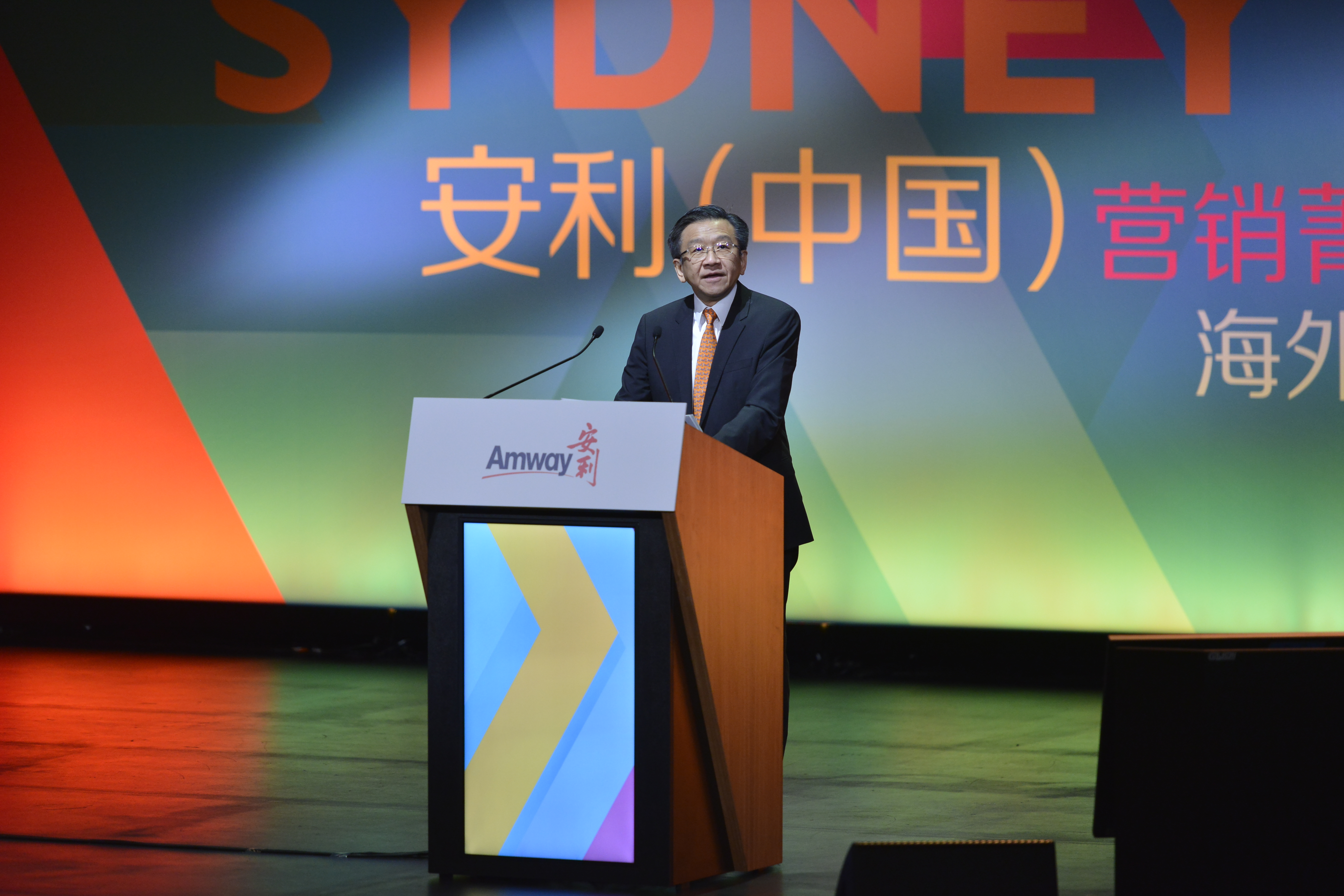 本次是安利（中国）营销菁英悉尼海外研讨会活动，身着正装的企业领导站在印有Amway安利标识的讲台后发言，背景大屏显示有“SYDNEY”“安利（中国）”“海外”等字样，现场氛围正式昂扬，彰显安利品牌活动的专业质感。