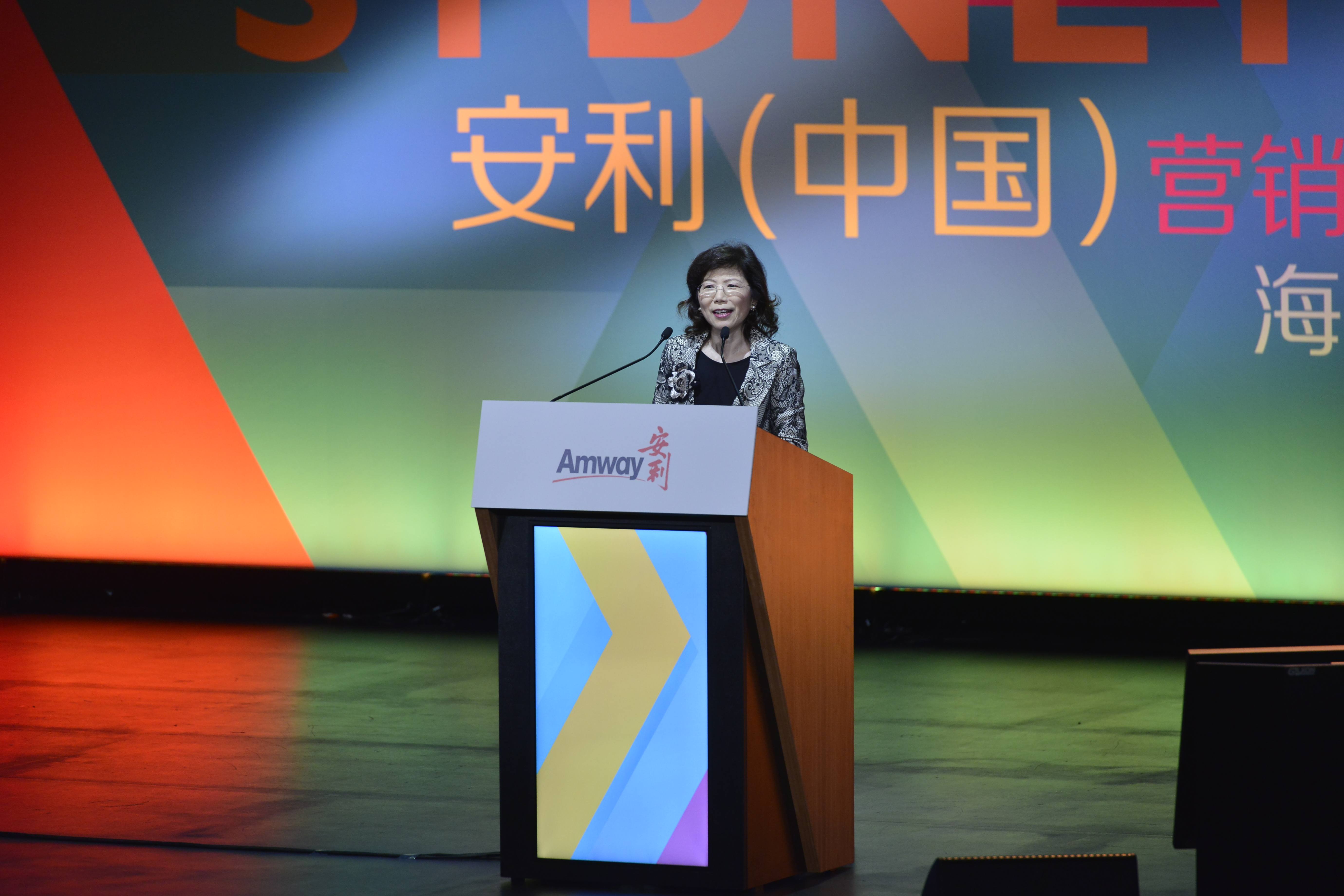 活动现场，女性嘉宾正站在印有“Amway 安利”标识的讲台后发言，背景大屏清晰显示“安利（中国）营销”字样，整体场景正式庄重，氛围积极明快，充分彰显安利品牌活动的专业质感与昂扬风貌。