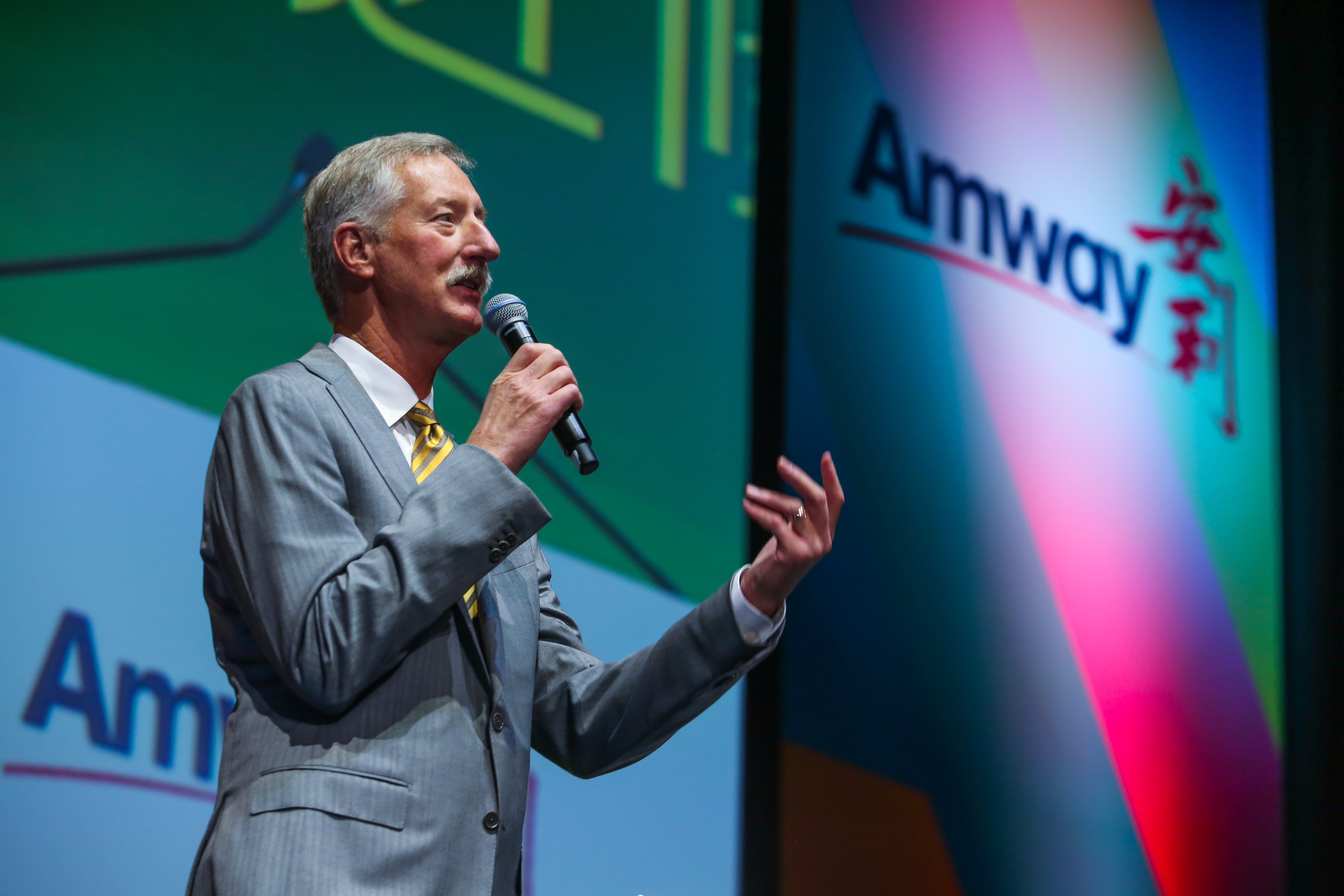 画面中身着灰色西装的外籍高管手持麦克风，站在印有「Amway」「安利」标识的活动舞台背景前发言，他神态从容、手势舒展，现场氛围正式热烈，充分展现出安利活动的专业质感与积极向上的品牌风貌。