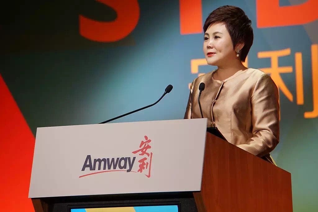 安利活动现场，身着香槟色质感套装的女性嘉宾站在印有“Amway 安利”标识的讲台后发言，台面配备双话筒，背景为活动主题大屏，整体氛围正式庄重，充分展现安利品牌活动的专业风貌。