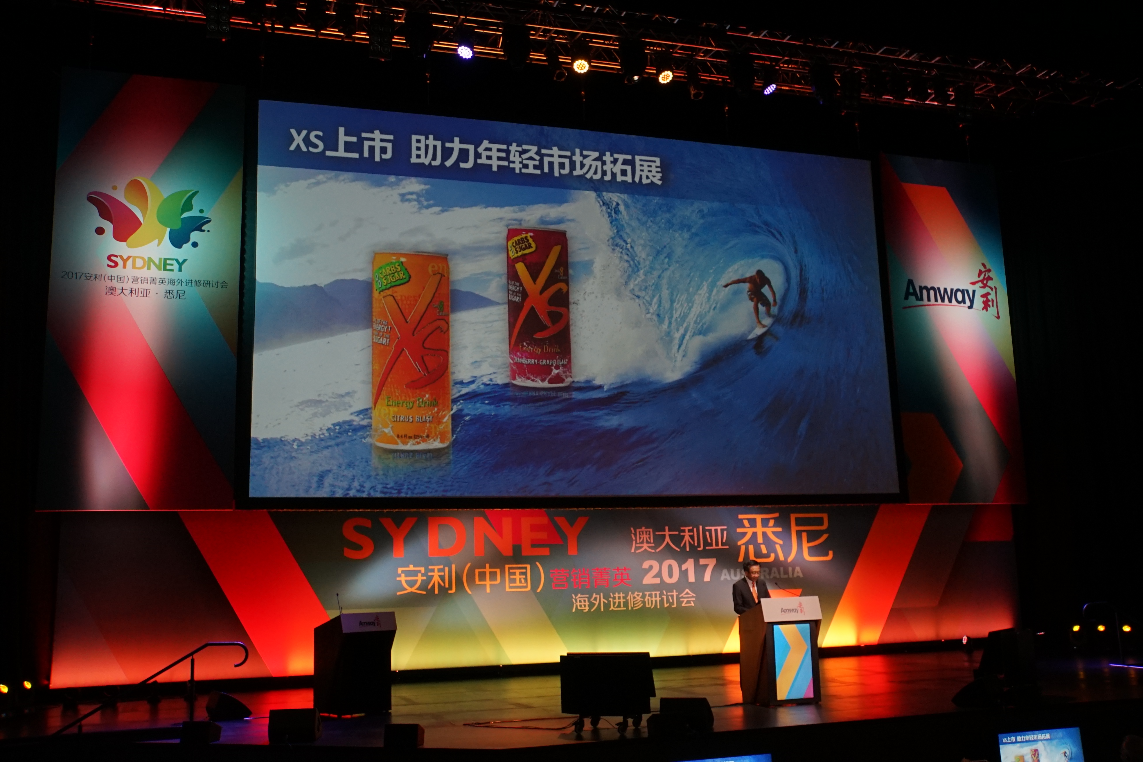 该场景为2017安利（中国）营销菁英澳大利亚悉尼海外进修研讨会现场，舞台大屏展示“XS上市 助力年轻市场拓展”内容，呈现两款XS能量饮料产品，演讲者在台前发言，现场氛围专业正式，体现安利布局年轻消费市场的规划。