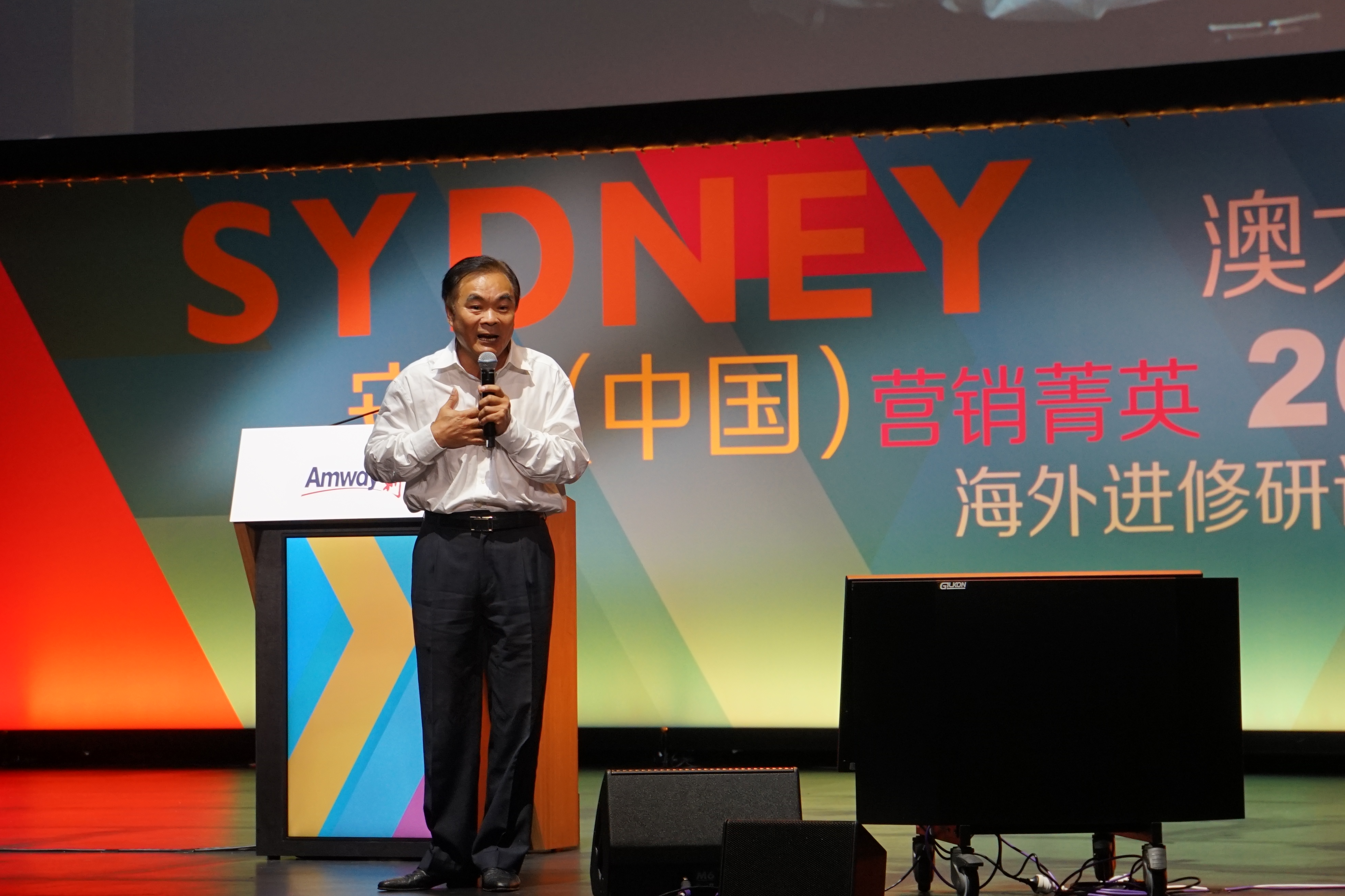 本场景为安利（中国）营销菁英悉尼海外进修研讨会现场，一位男士站在印有Amway标识的讲台旁持话筒致辞，背景大屏标注“SYDNEY”“营销菁英海外进修研”等活动字样，氛围庄重积极，彰显安利对营销伙伴的成长支持。