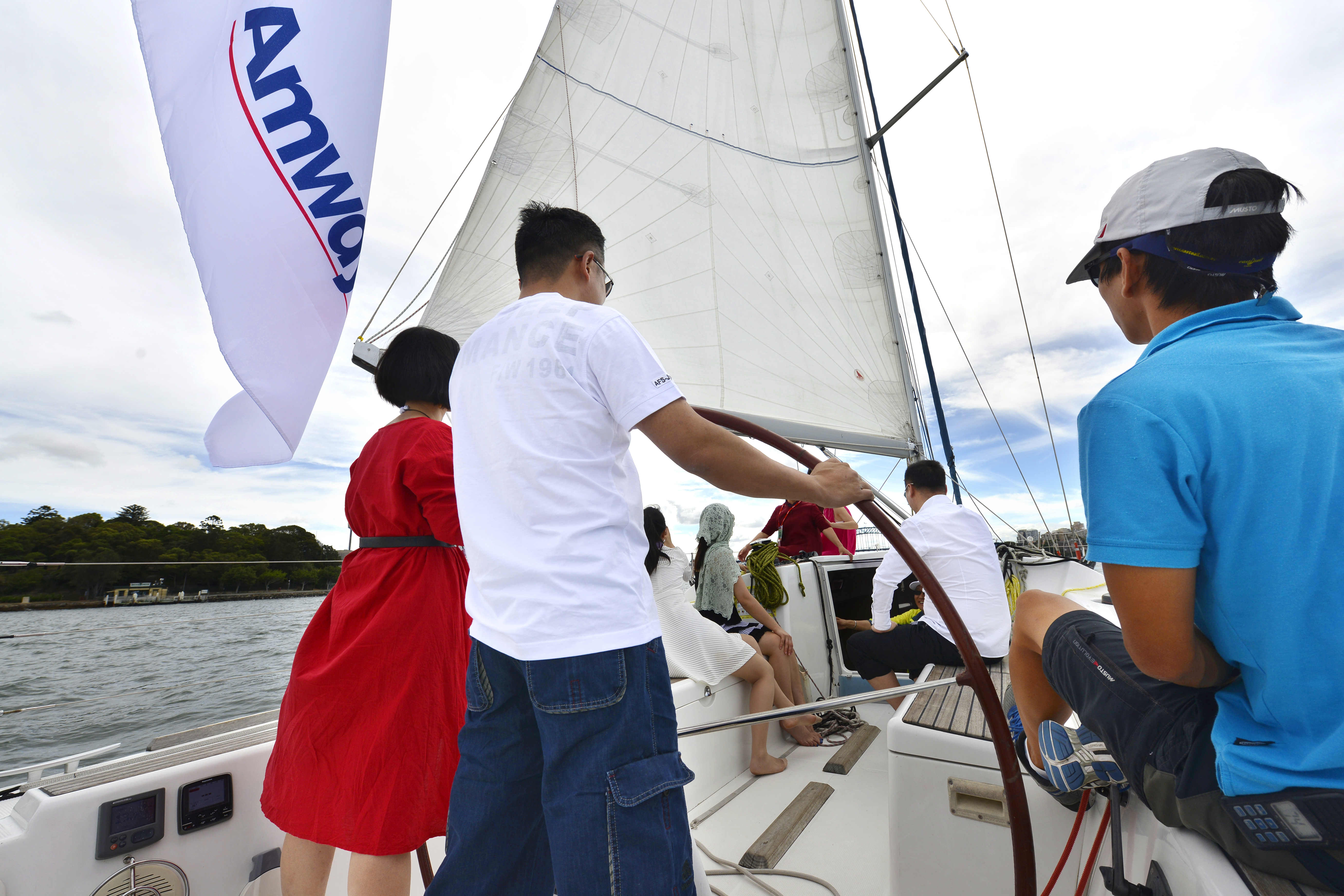 帆船上悬挂着印有“amway”字样的安利品牌旗帜，多名安利伙伴共同参与帆船航行体验，有人掌舵把控航向，其余伙伴或站或坐于船身，海面开阔清朗，整体氛围轻松融洽，尽显团队同心前行的蓬勃朝气，契合安利积极向上的品牌调性。