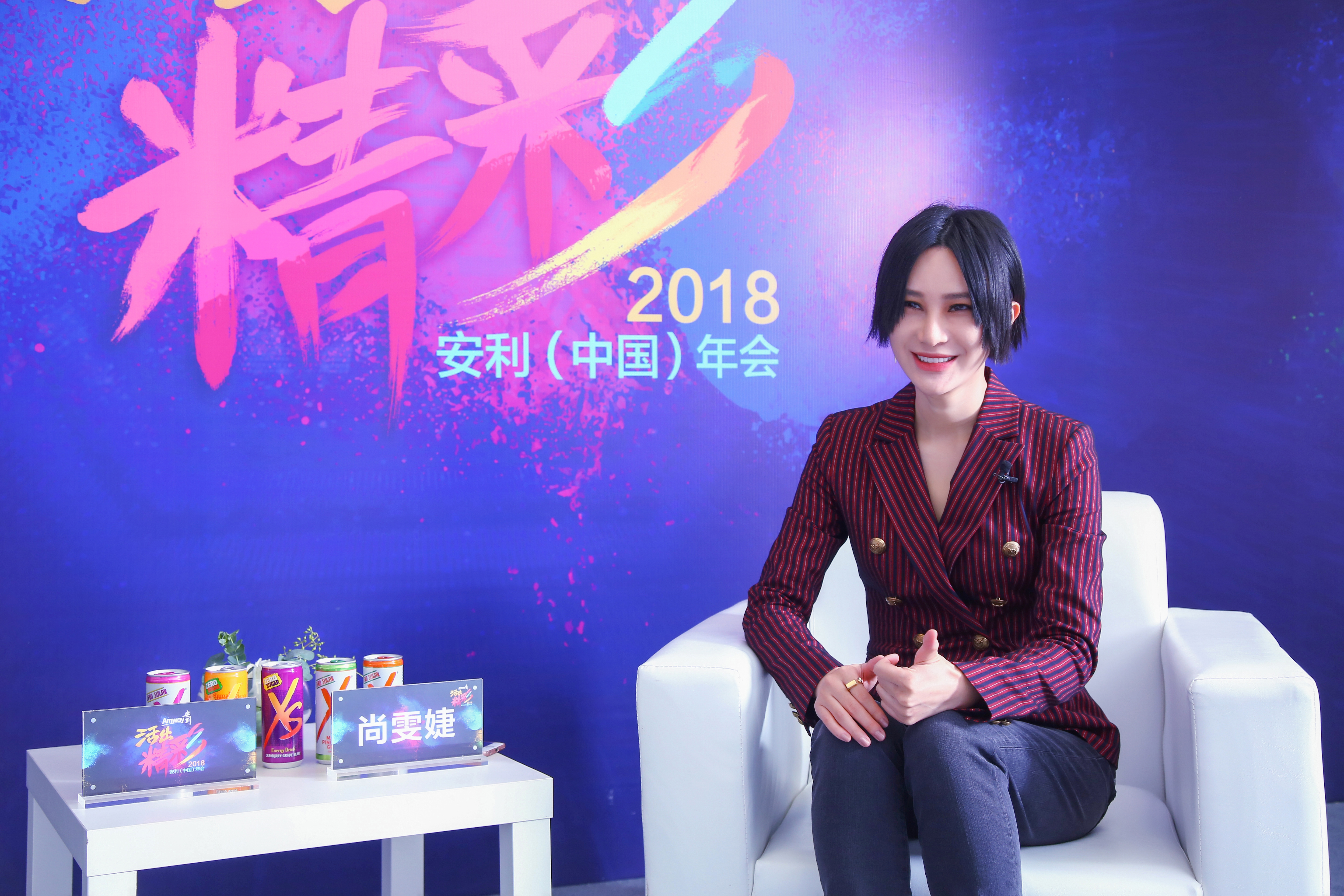 本次为2018安利（中国）年会访谈现场，特邀嘉宾尚雯婕坐于白色沙发上笑容亲和。面前白桌摆放XS系列饮品、印有“活出精彩”的活动主题牌与尚雯婕姓名牌，背景为对应年会主题的蓝紫色背景板，整体氛围明快活力，尽显年会积极向上的氛围。