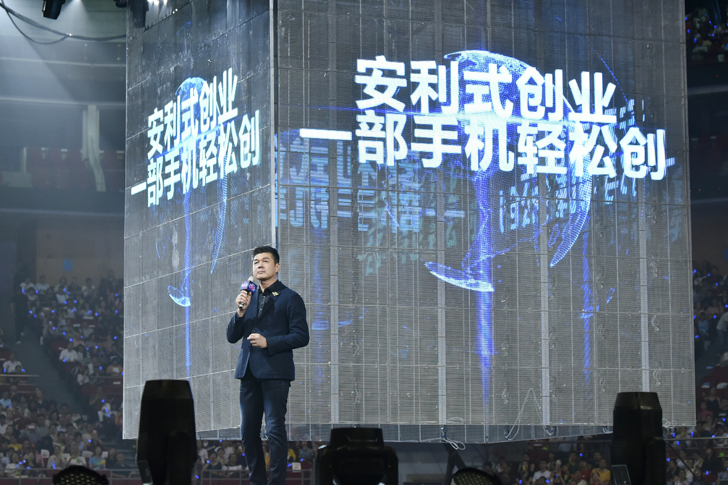 活动现场，主讲人手持话筒站在舞台上进行分享，身后透明大屏清晰呈现“安利式创业 一部手机轻松创”的核心主题，台下座无虚席，氛围热烈庄重，充分展现安利轻量化创业的发展路径，为在场创业者传递清晰的事业方向。