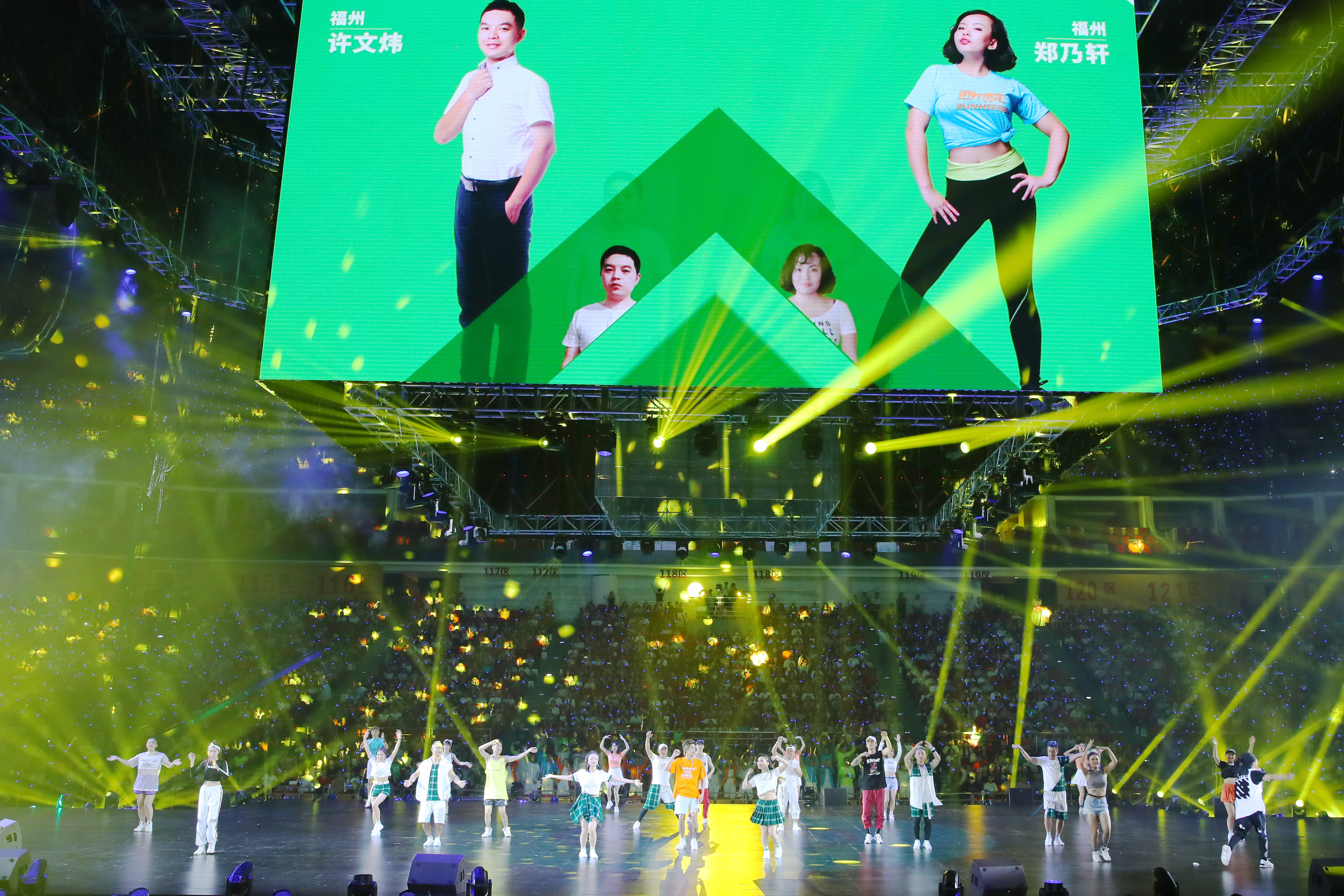 本次安利福州年会现场灯光炫目，舞台上多位表演者活力起舞，上方巨幅屏幕展示福州优秀伙伴许文炜、郑乃轩的肖像，台下座无虚席，全场氛围热烈昂扬，尽显安利品牌的活力与团队向心力。