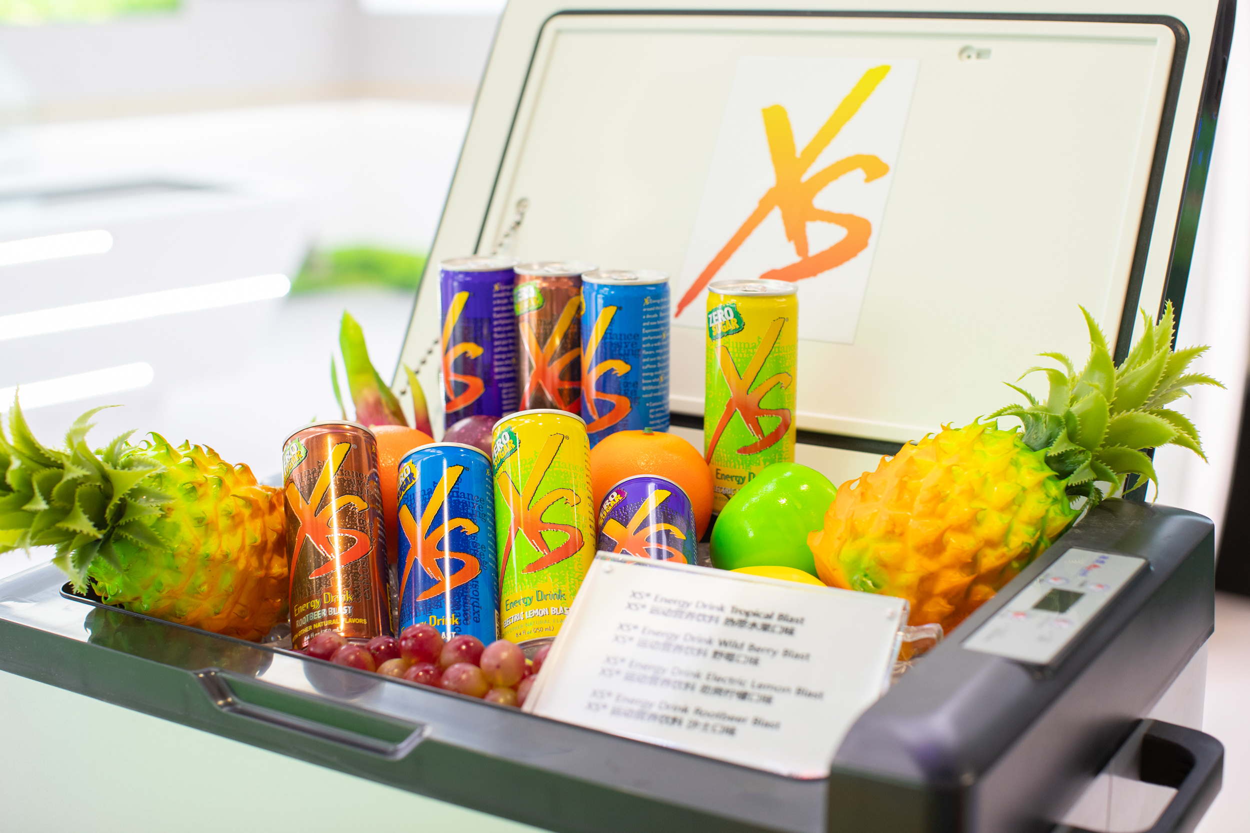 画面为安利XS运动营养饮料陈列场景，印有XS标识的冷藏柜前，摆放着多口味XS运动饮料，搭配菠萝、橙子、葡萄等新鲜鲜果，立牌标注了热带水果、野莓、劲爽柠檬、沙士等多款口味，整体氛围鲜活明快，凸显产品健康活力的特质。