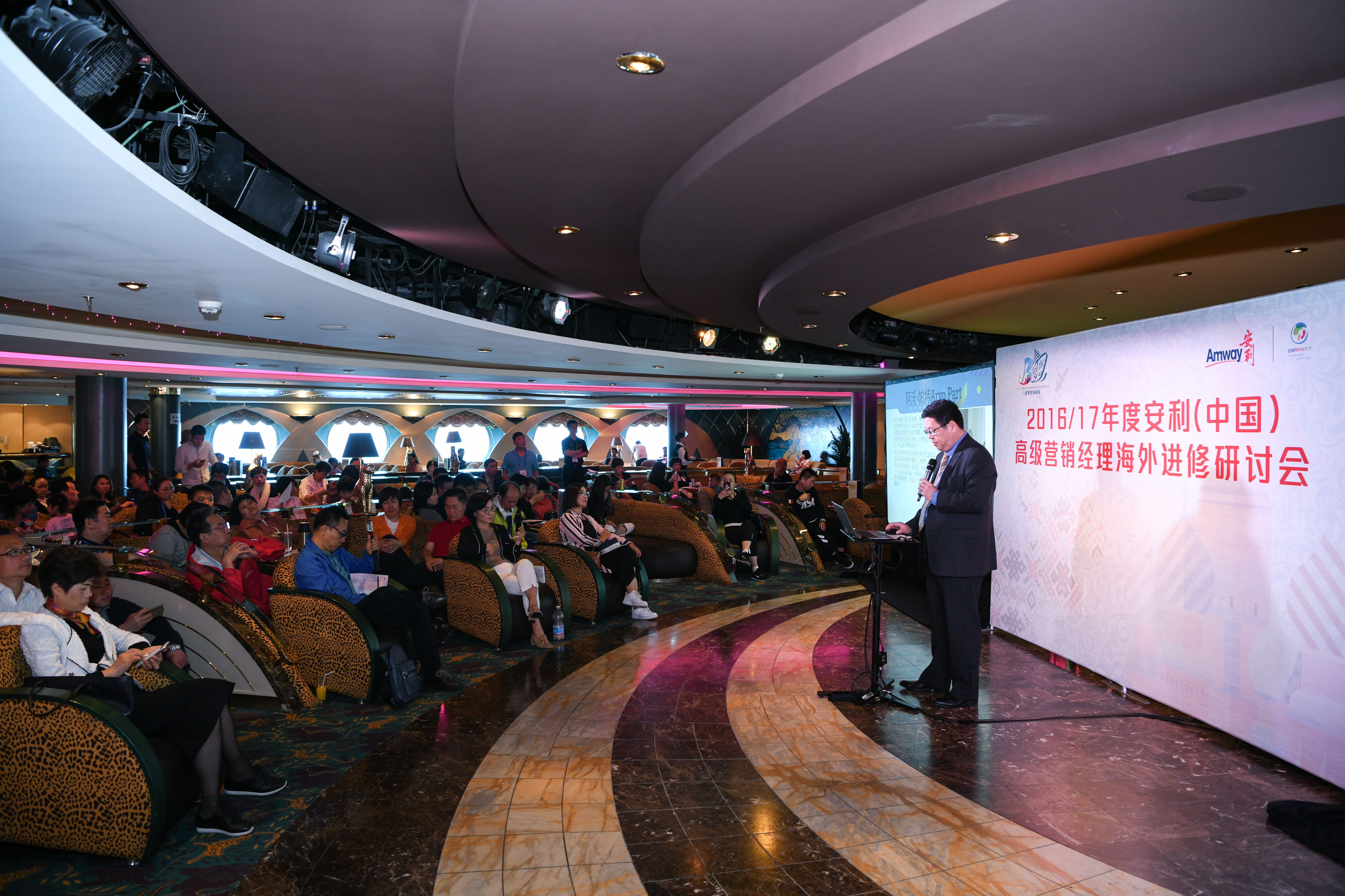 这是2016/17年度安利（中国）高级营销经理海外进修研讨会现场，身着正装的讲师持麦在台前开展分享，台下众多营销经理端坐认真聆听，会场氛围专注热烈，充分展现出安利伙伴积极进取、共促成长的良好风貌。