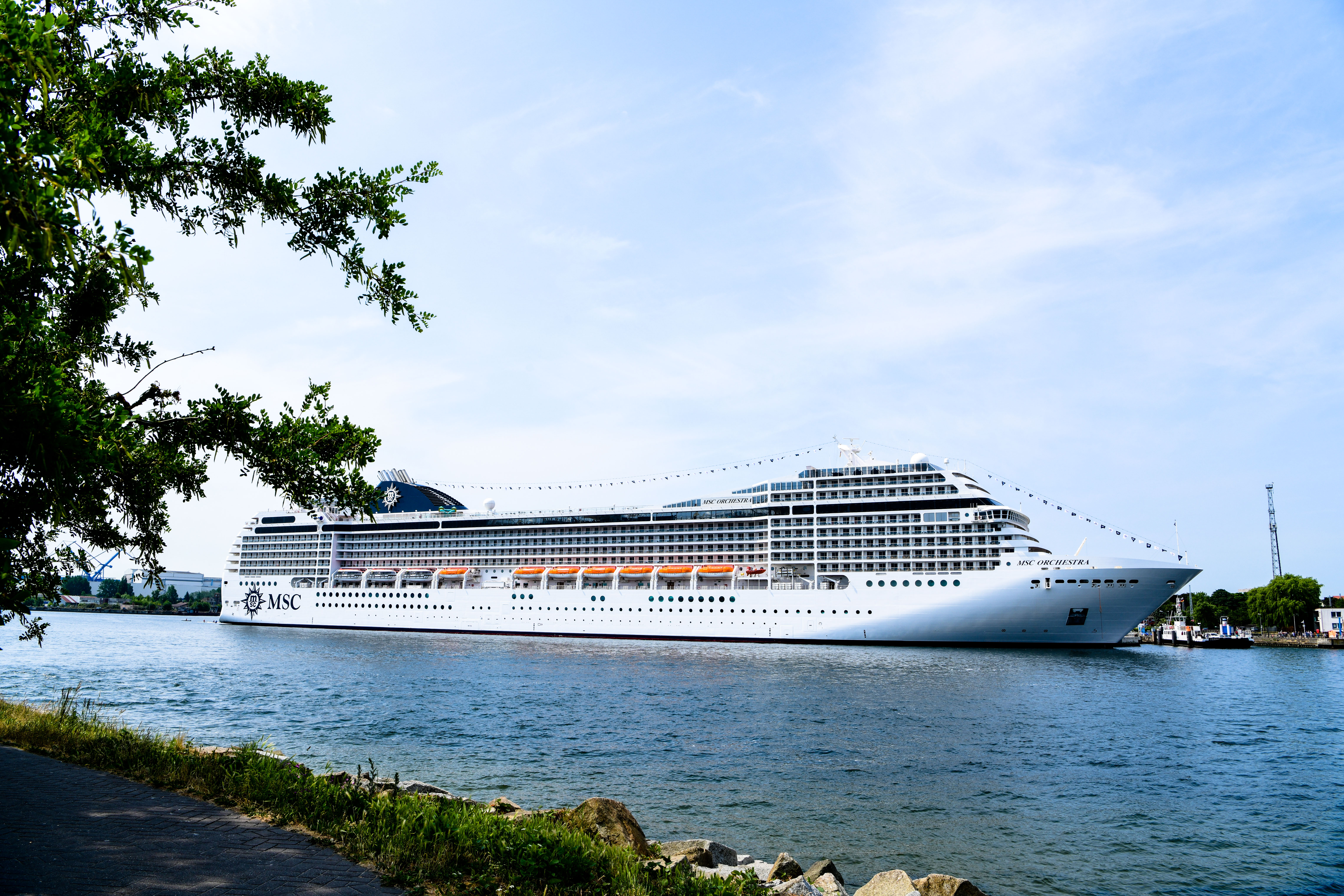 澄澈晴空下，碧波荡漾的港湾中，印有“MSC”标识的MSC ORCHESTRA豪华邮轮稳稳停泊，船体洁白恢弘，岸边绿植掩映景致清新，整体氛围开阔明快，彰显安利为伙伴打造的高端尊享旅程即将开启，尽显品牌雄厚实力与暖心关怀。