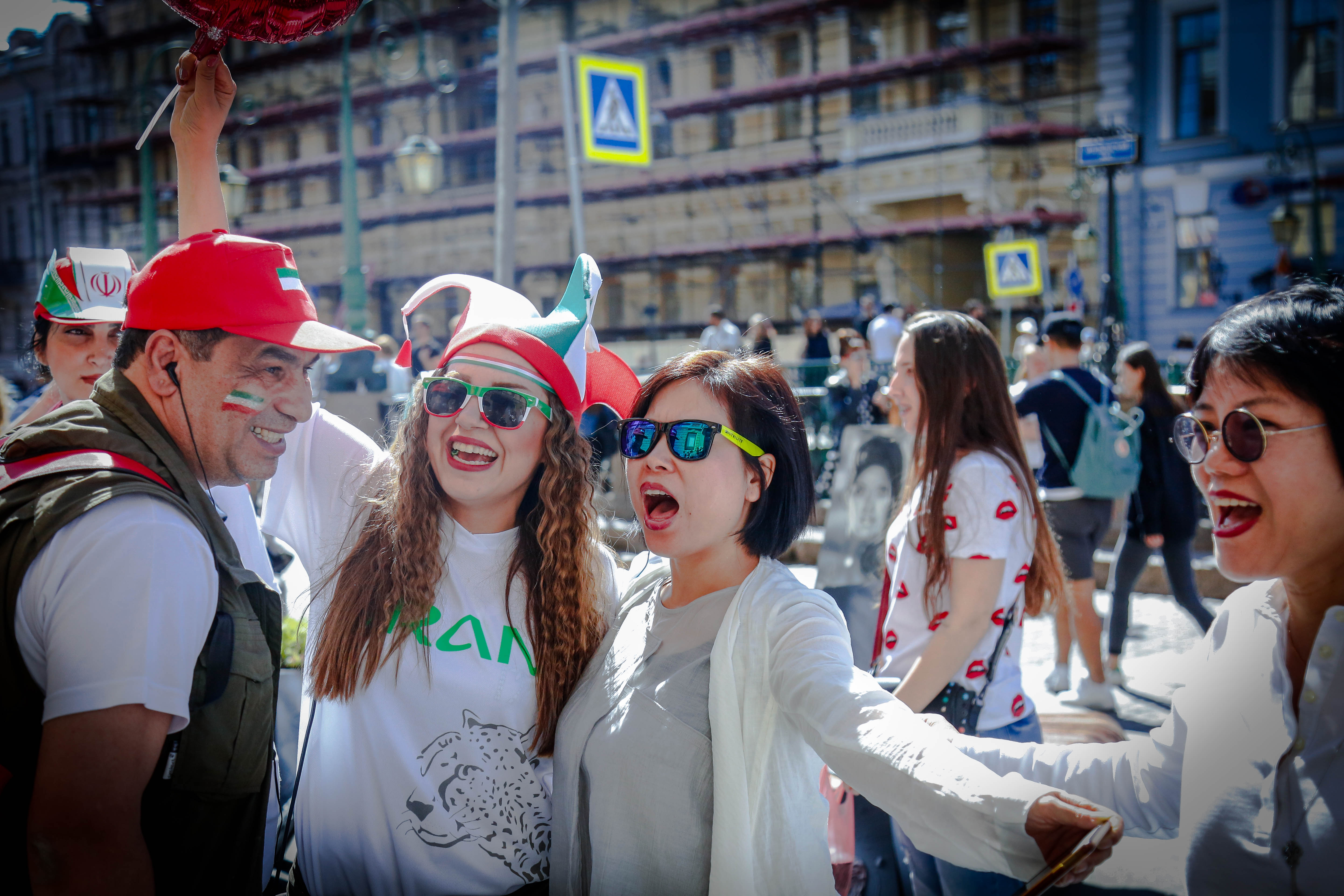 安利海外交流活动现场，伙伴们与身穿印有“IRAN”字样服饰、佩戴伊朗主题特色装饰的当地友人在异国街头亲密同框，众人脸上满是开怀笑容，氛围轻松热烈，尽显不同国籍伙伴融洽相处的温暖氛围，传递出积极欢快的活动气质。