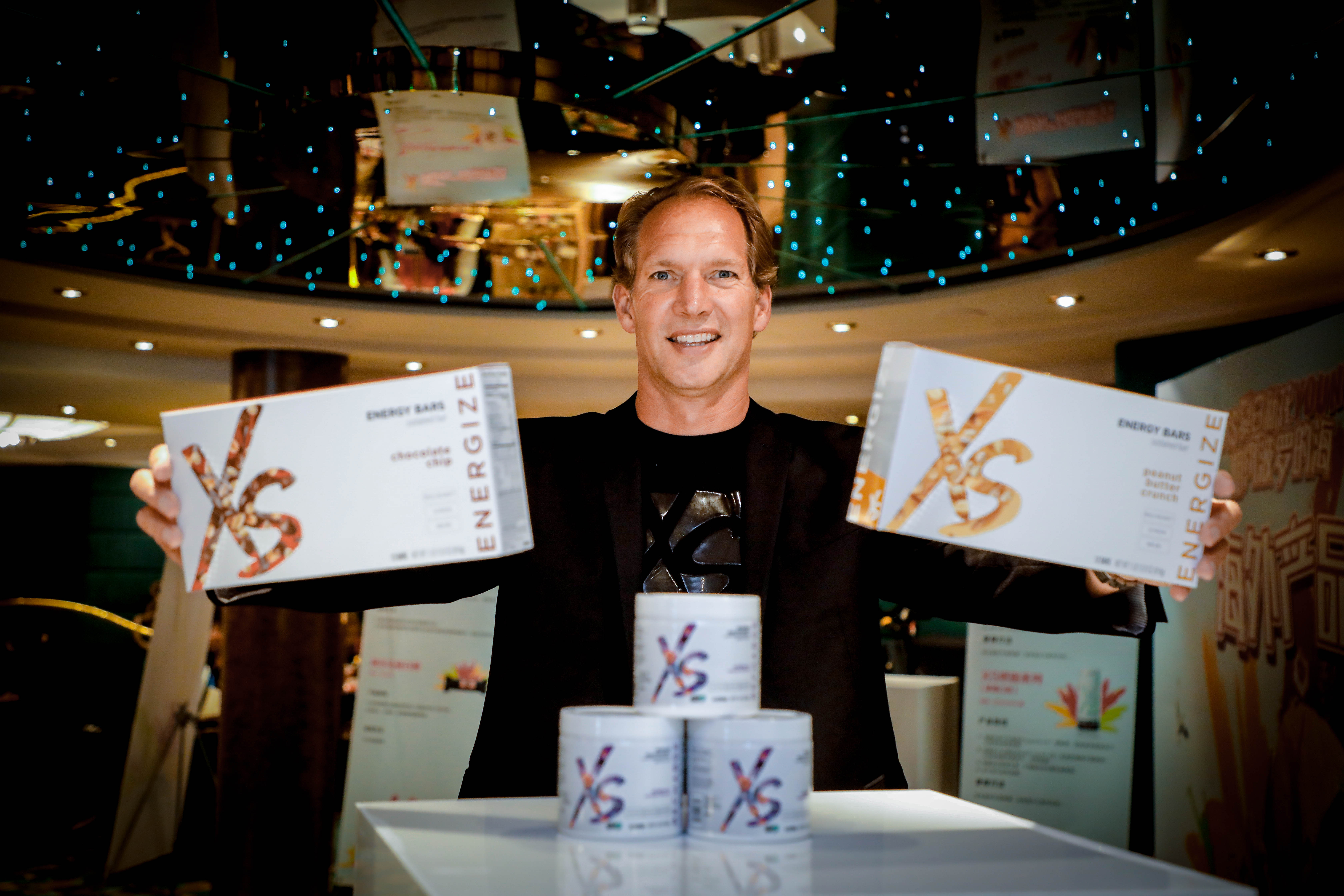 活动现场，外籍嘉宾面带笑容，双手分别展示印有“XS”“ENERGIZE”“ENERGY BARS”标识的巧克力味、花生酱味XS能量棒，身前展台上摆放着三罐XS系列营养产品。会场布置精致明亮，氛围积极专业，凸显出安利XS运动营养产品线的活力特质。