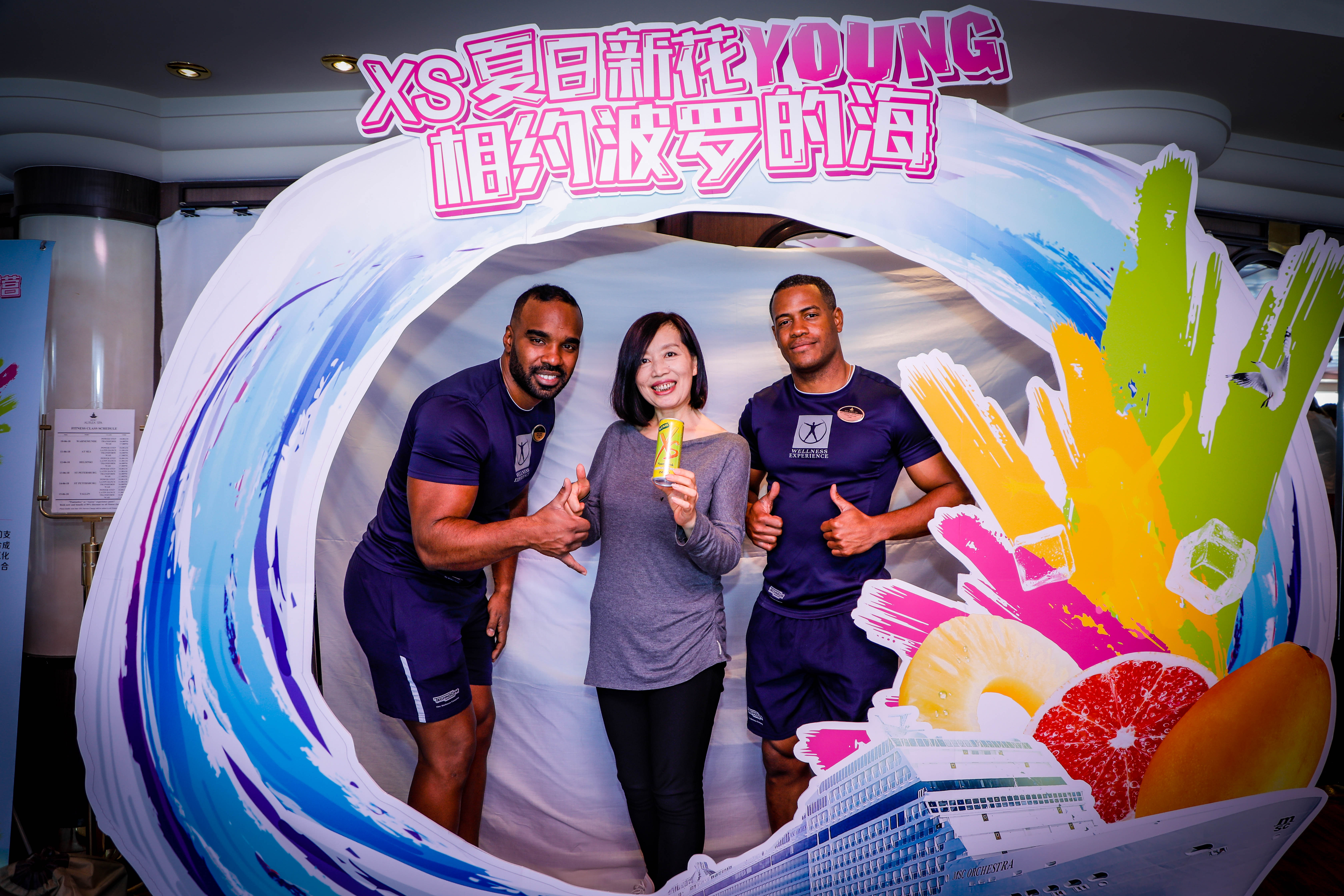 活动打卡装置印有“XS夏日新花YOUNG 相约波罗的海”主题字样，一名女士手持XS饮料，与两位身着运动服的男士在装置内合影，三人面带笑容、比出点赞手势，现场氛围轻松欢快，尽显活动活力年轻的基调。
