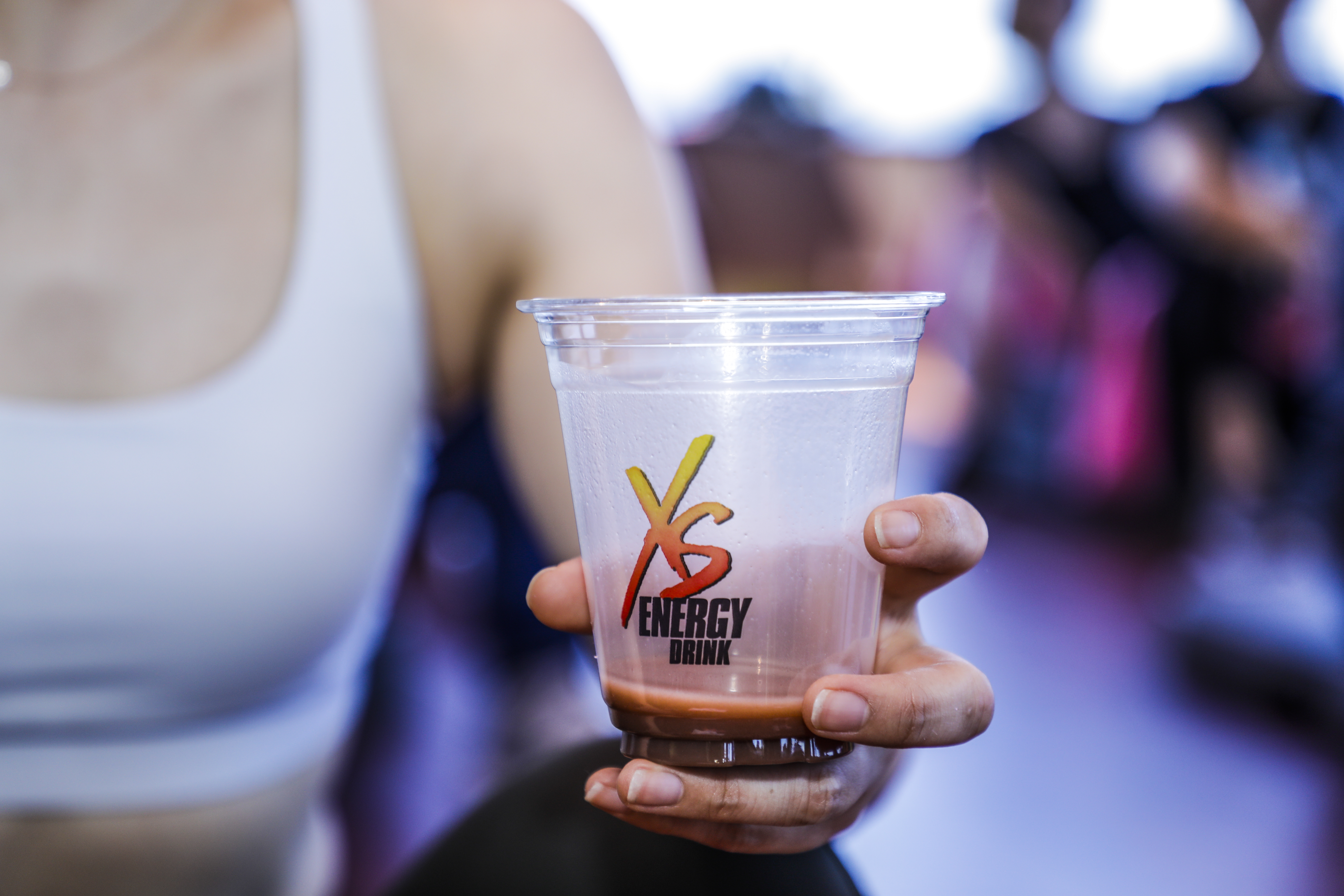 画面中一只手握持印有「XS ENERGY DRINK」标识的透明杯装XS能量饮品，杯身凝着的水珠尽显冰爽质感，背景为模糊的安利活动现场，整体氛围鲜活有活力，传递出安利健康活力的品牌理念，为参与者补足能量。
