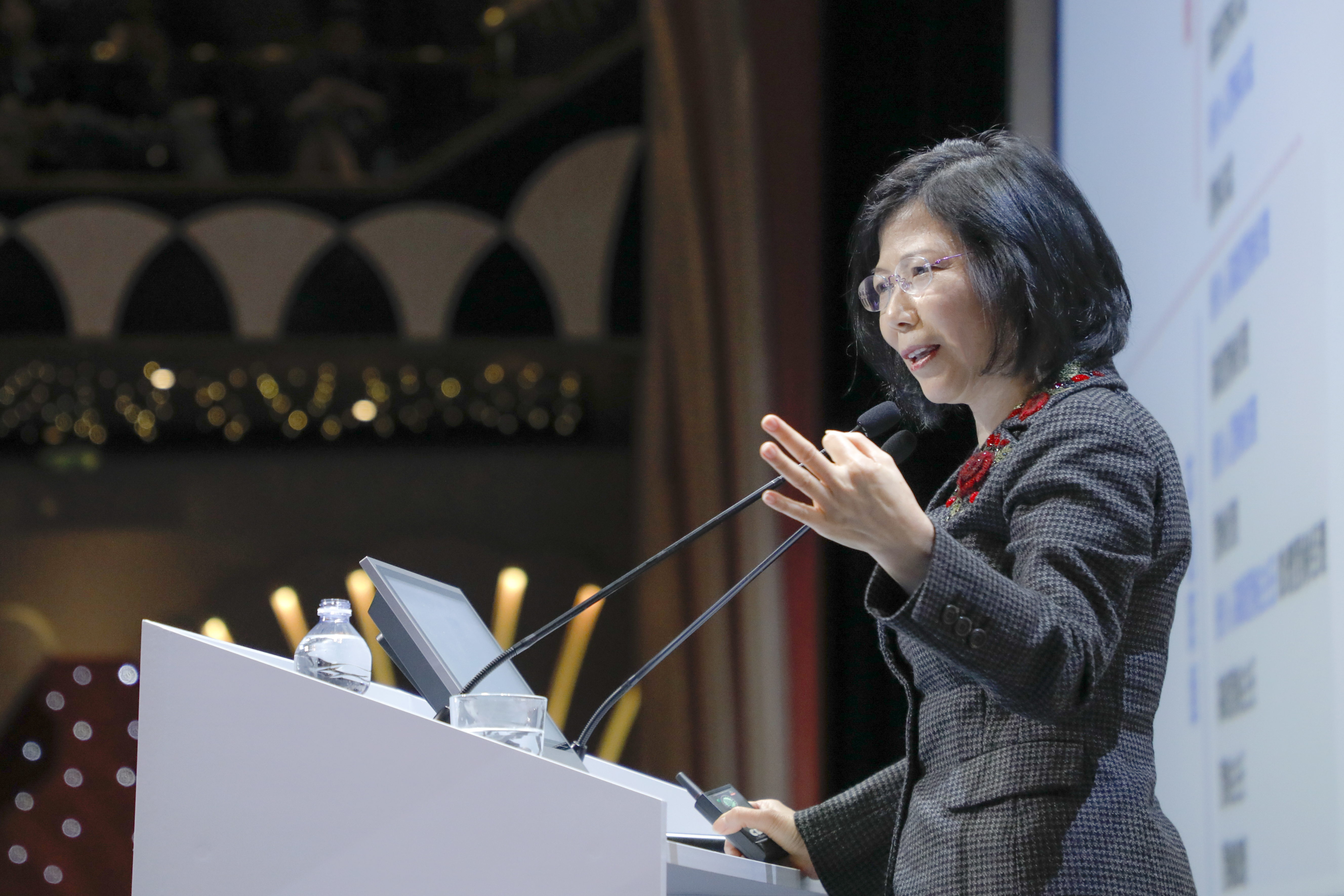 安利研讨会活动现场，女性嘉宾站在白色演讲台前，手持翻页器对着麦克风进行主旨分享，手势舒展从容，表述专业清晰。讲台上摆放着饮用水与演示屏，现场氛围庄重专业，传递出活动务实向上的积极基调。