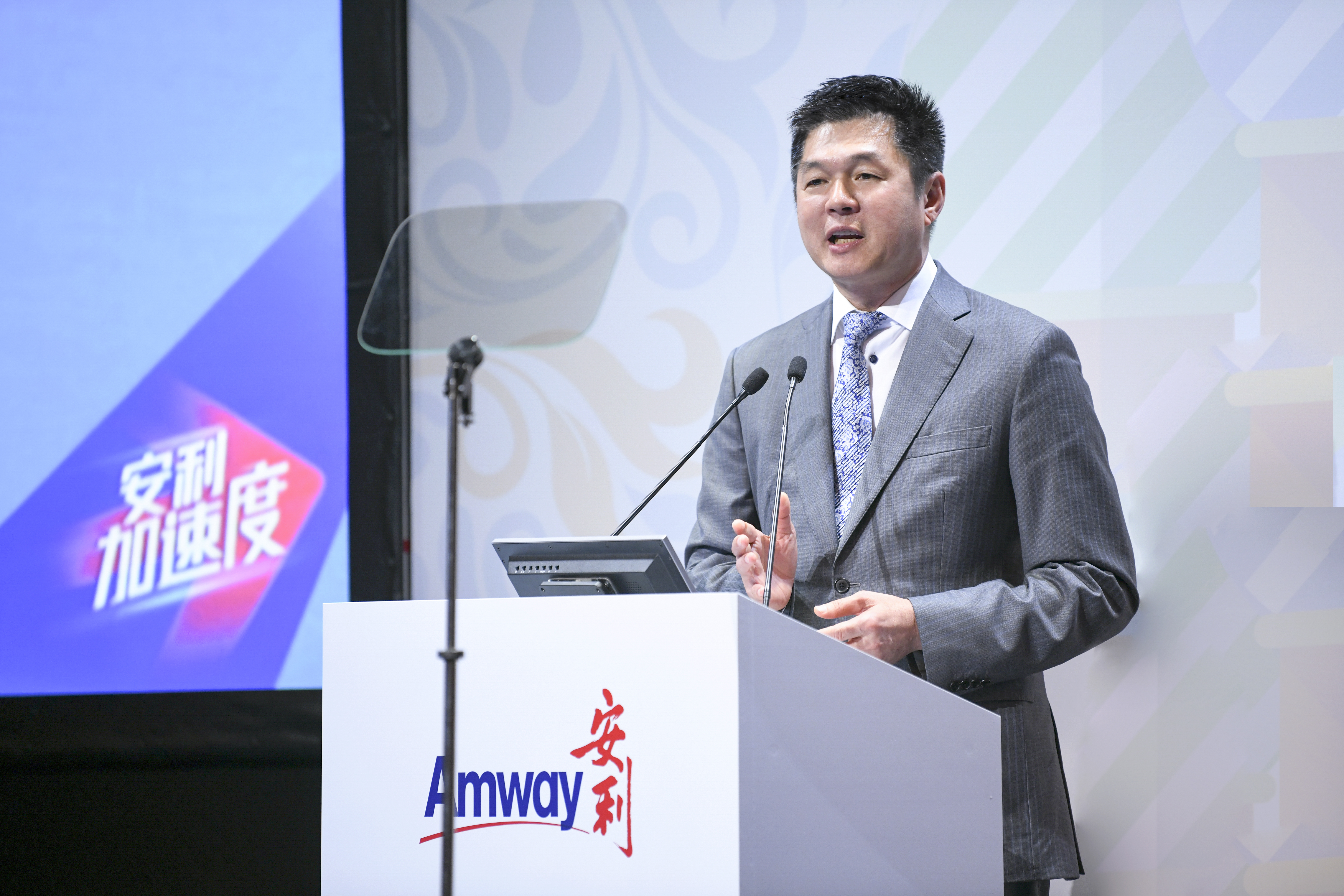 活动现场，身着灰色西装的高管站在印有“Amway 安利”标识的讲台后发言，背景大屏醒目展示“安利加速度”字样。高管发言状态昂扬，现场氛围正式积极，充分传递出安利锐意进取的发展导向。