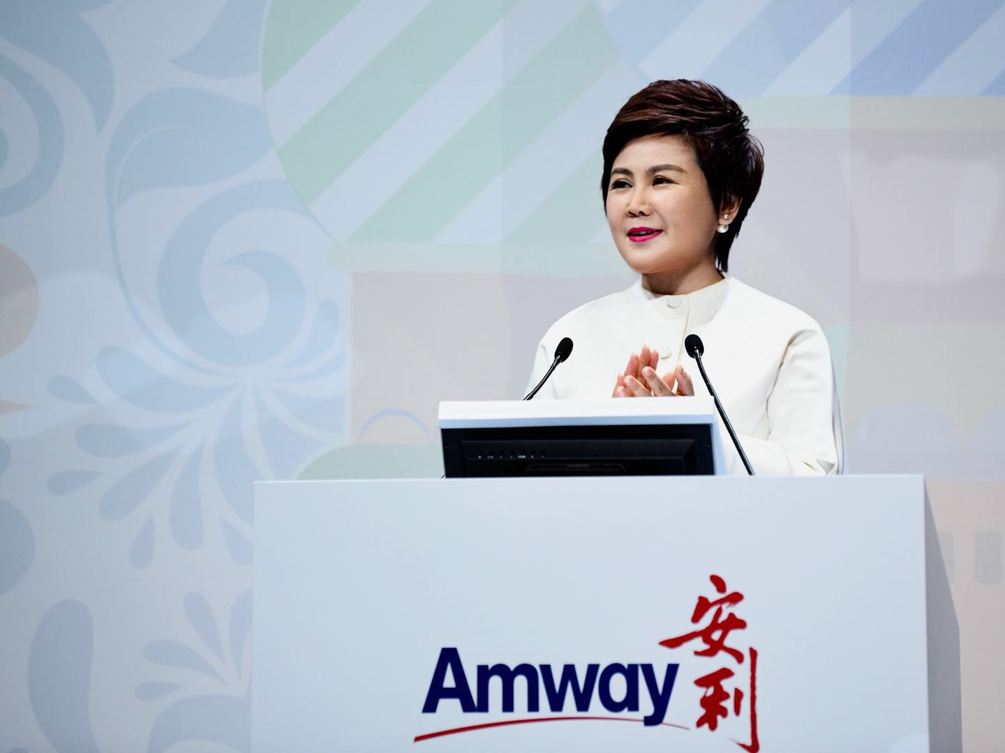 画面中身着白色正装的女士站在印有“Amway 安利”标识的讲台后，面带微笑，双手抬起作鼓掌姿态，背景为带有柔和纹样的活动背景板，现场氛围正式积极，尽显安利品牌活动的专业风貌。