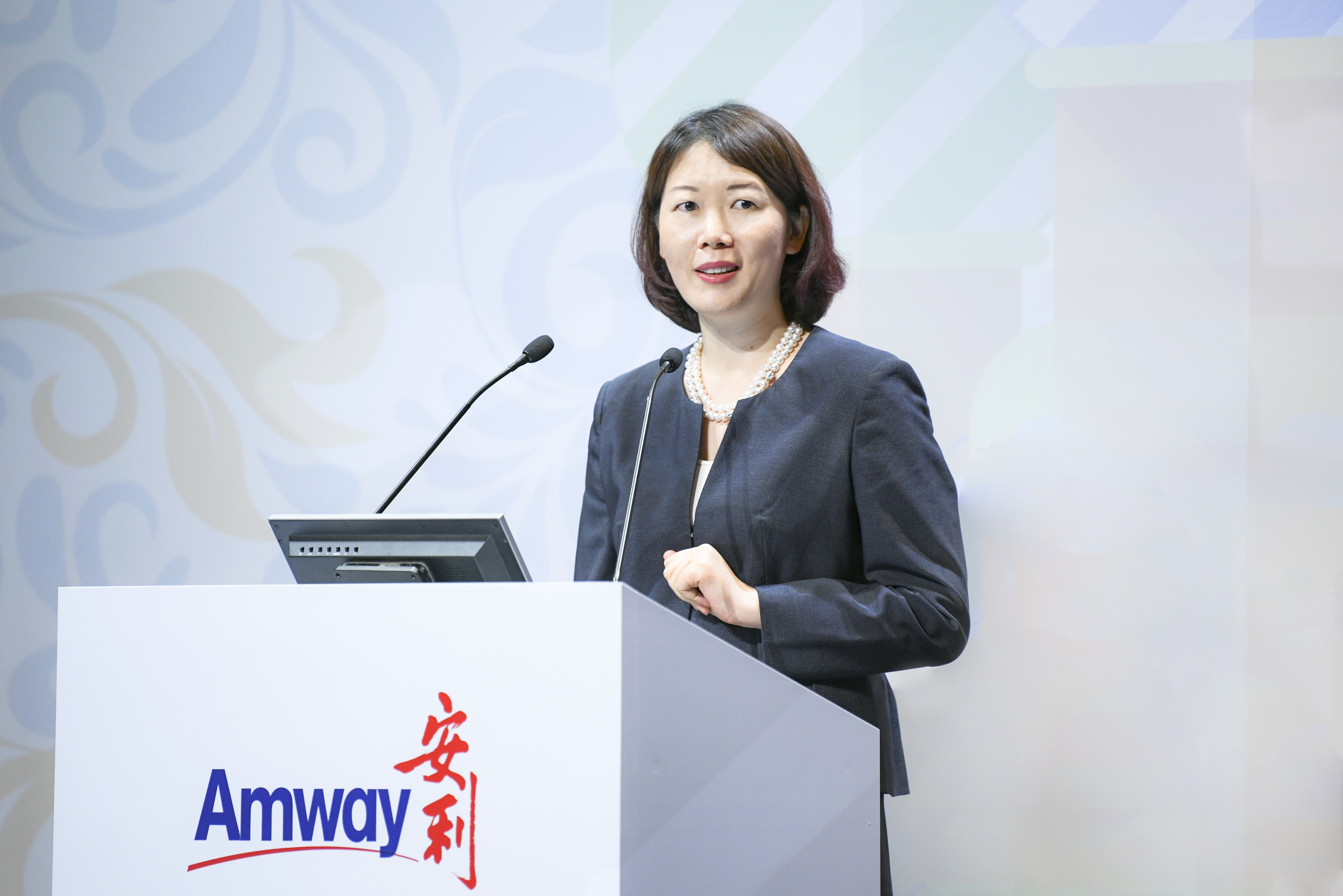 画面中，身着深色正装、佩戴珍珠项链的女性高管站在印有“Amway 安利”标识的讲台后发言，她神态从容专业，现场氛围正式积极，充分彰显安利品牌活动的专业质感与正向风貌。