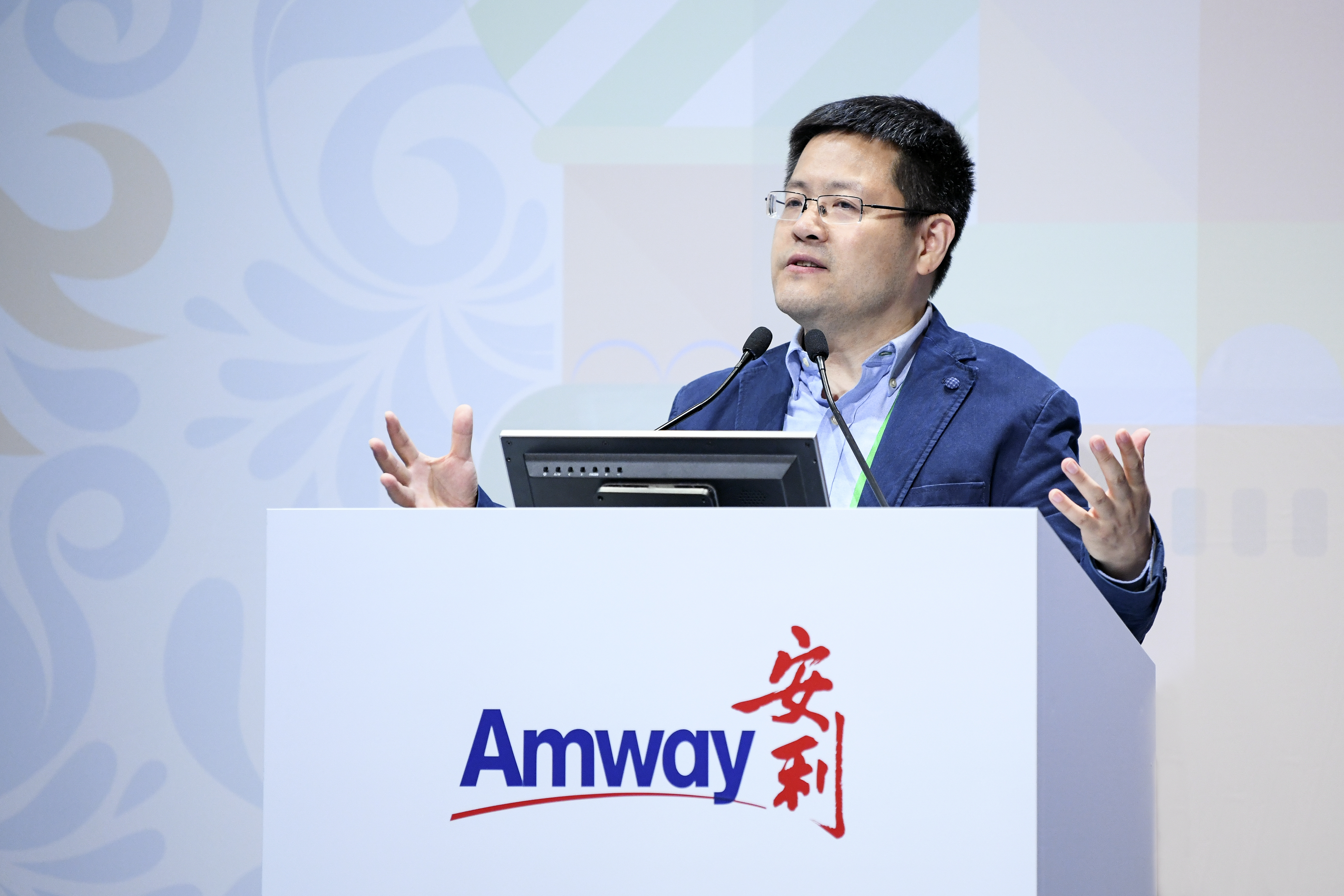 安利活动现场，嘉宾站在印有“Amway”“安利”标识的演讲台后，身着蓝色外套，正张开双手投入发言，现场氛围专业正式，传递出积极向上的交流氛围，尽显安利品牌活动的专业调性。