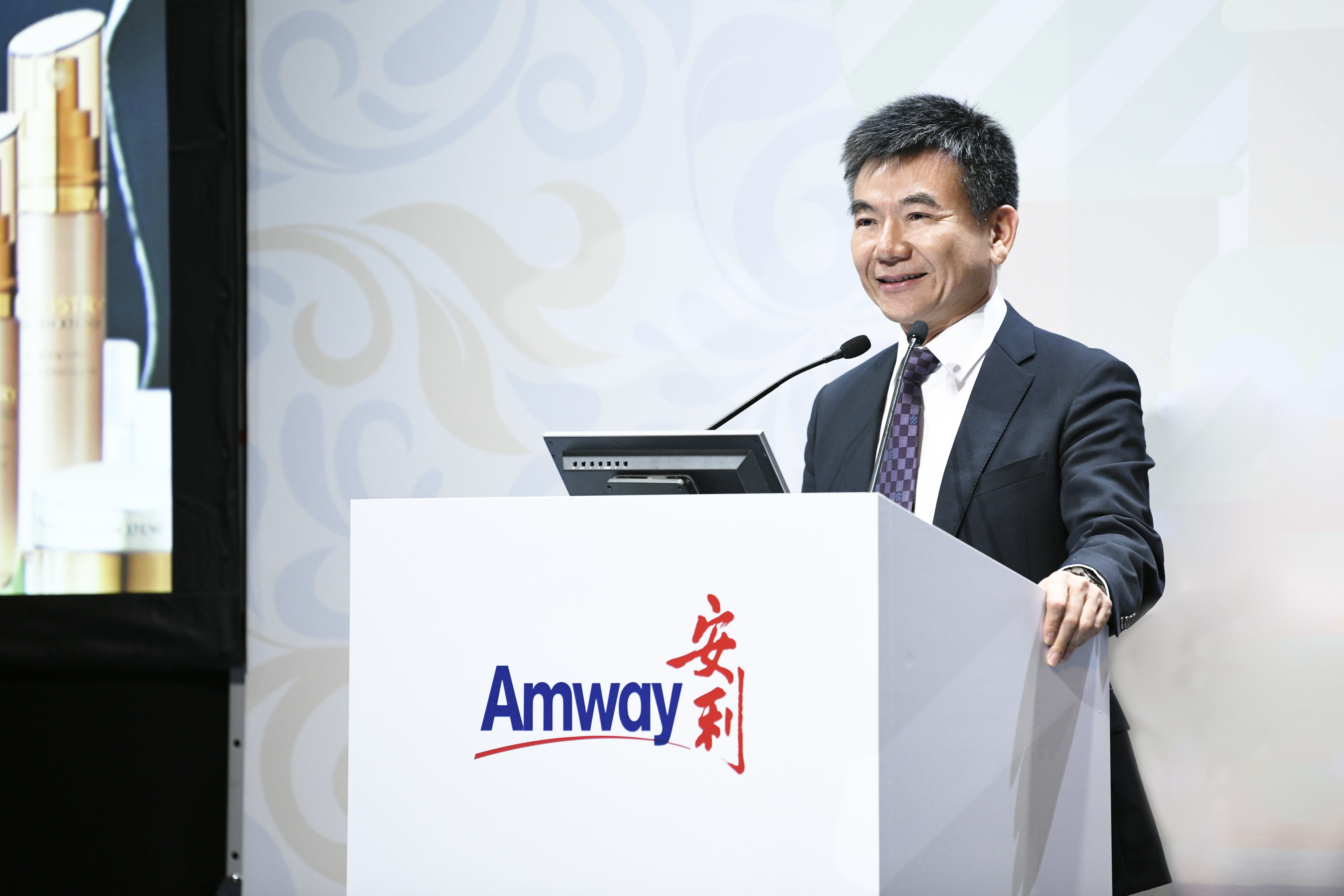 活动现场，身着正装的领导站在印有“Amway 安利”标识的讲台后发言，面带笑意、神态从容，背景侧边展示安利护肤产品，现场氛围庄重专业，传递出安利积极正向的品牌气质，契合企业正式活动的专业调性。