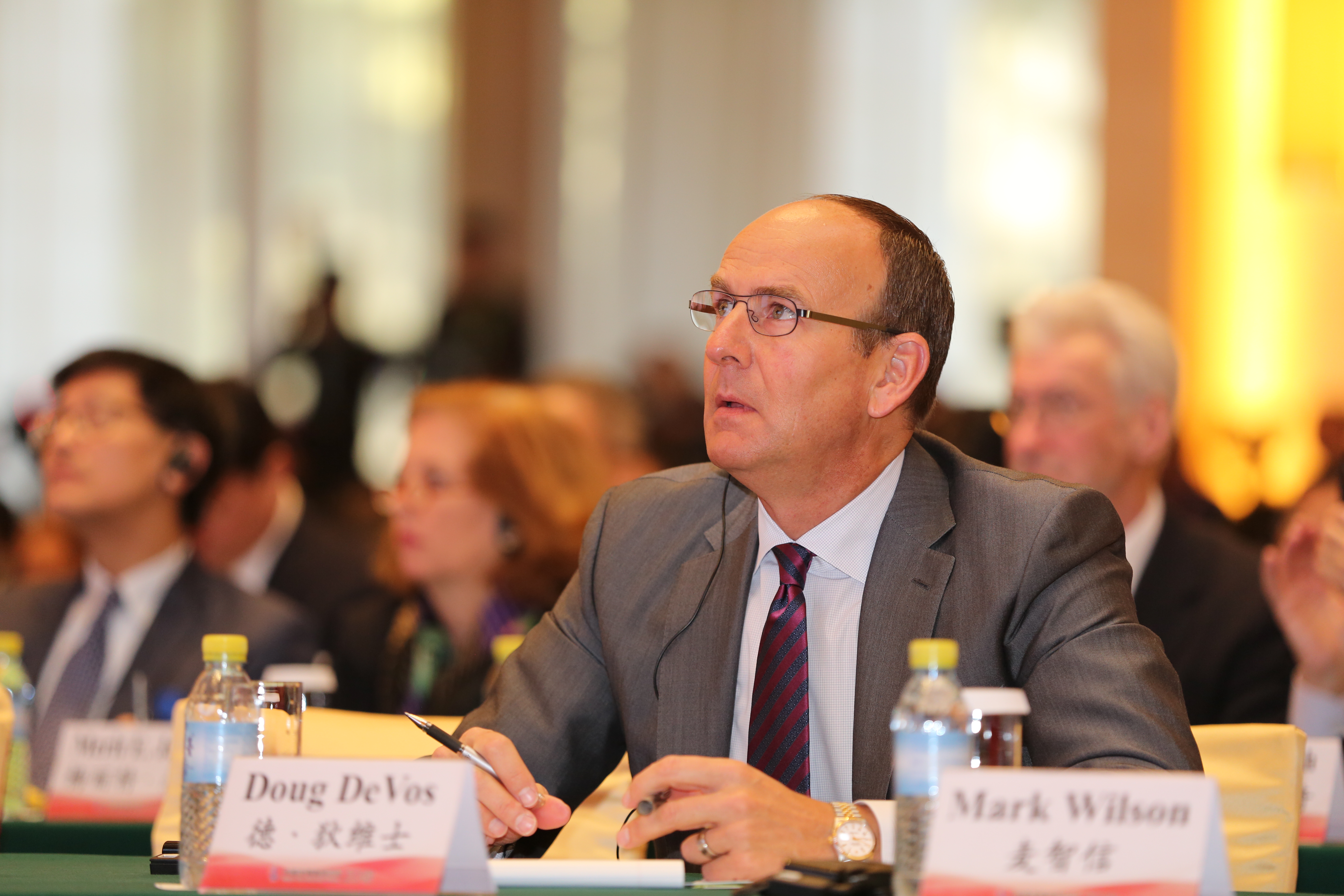 画面中安利高层德·狄维士（Doug DeVos）身着正装端坐会议桌前，手持笔专注聆听会议内容，桌面摆放着标注其姓名与麦智信（Mark Wilson）姓名的席卡，会场内还有多位参会嘉宾，氛围专业庄重，尽显会议的高规格正式感。