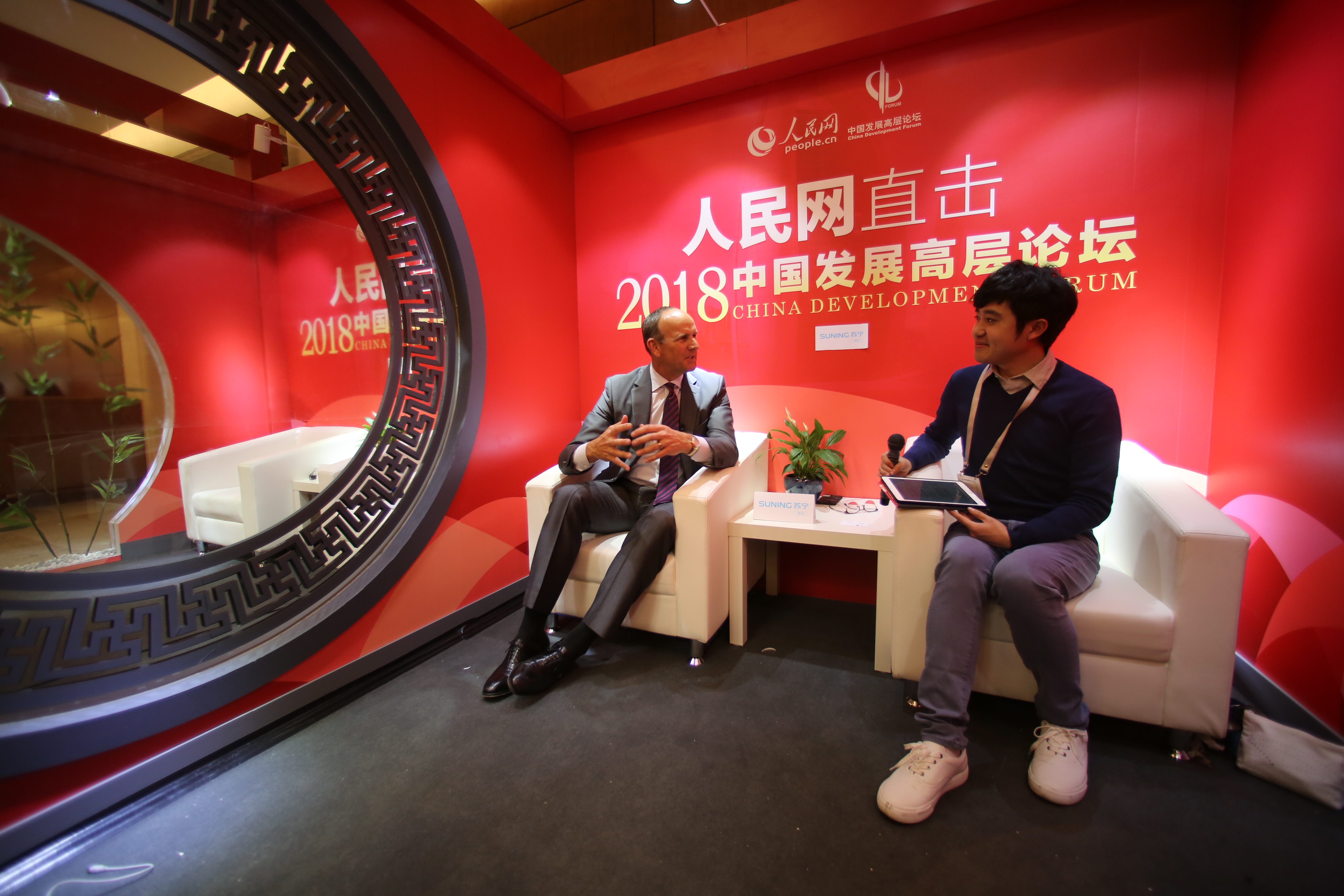 背景为印有“人民网 直击2018中国发展高层论坛”字样的红色主题墙，访谈间搭配中式回纹圆窗，典雅大气。外籍受访嘉宾与持采访设备的记者相对而坐，桌台摆放苏宁标识牌，二人交流顺畅，氛围专业融洽，尽显论坛深度对话的良好风貌。