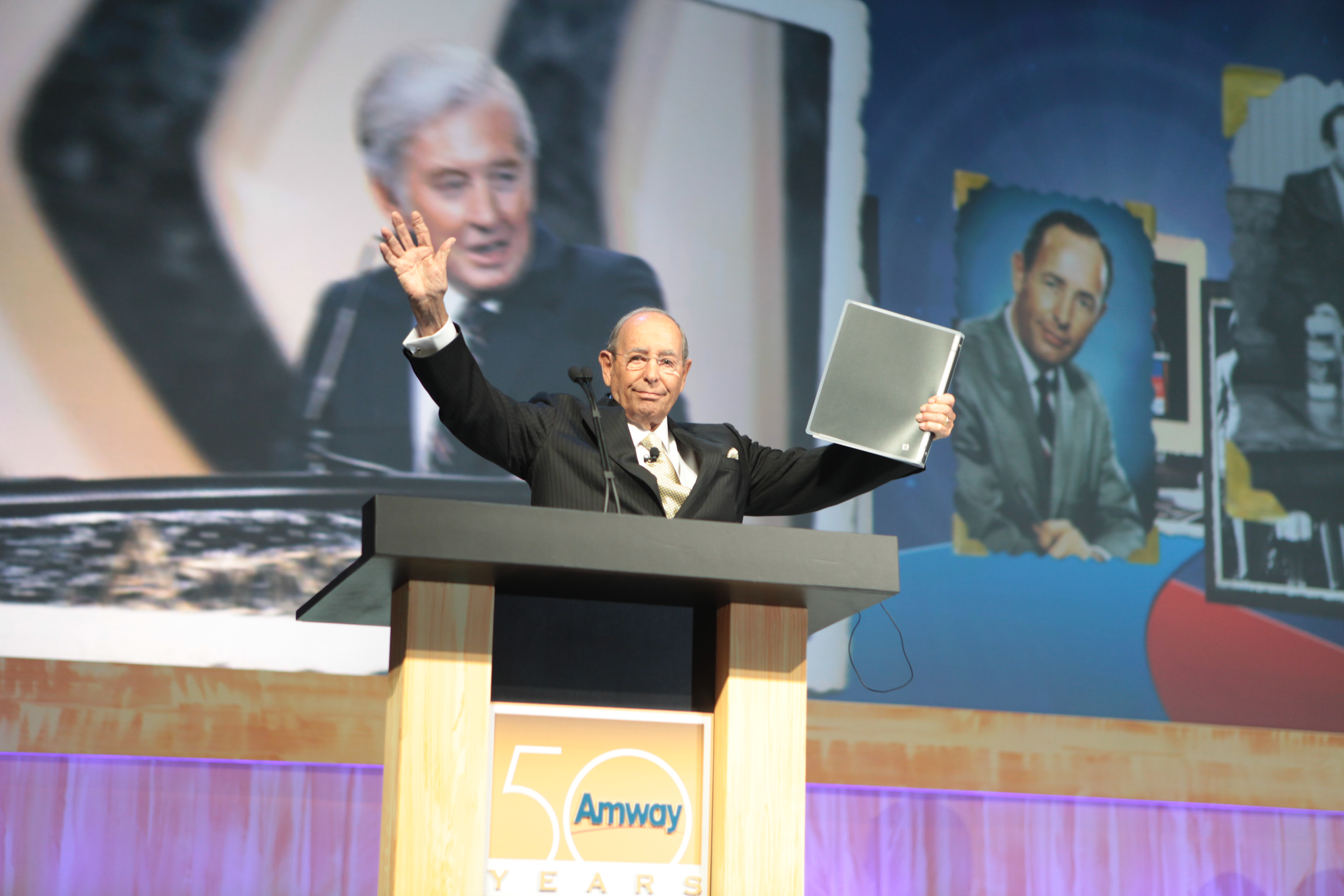 画面为安利50周年庆典活动现场，讲台印有核心标识“Amway 50 YEARS”，身着正装的嘉宾站在讲台后，一手挥手向台下致意，一手持文件夹，背景大屏展示安利发展相关历史影像，现场氛围庄重热烈，尽显品牌深厚底蕴与昂扬风貌。
