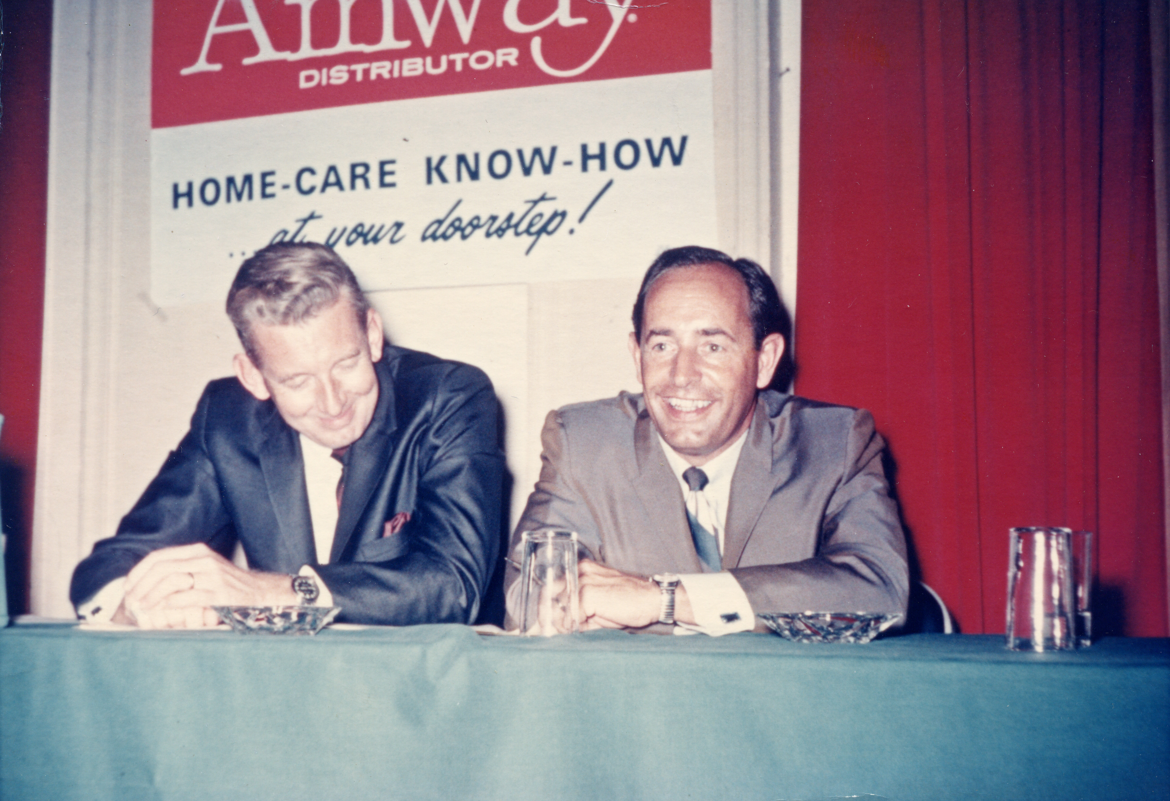 现场背景标识印有“Amway DISTRIBUTOR”“HOME-CARE KNOW-HOW ...at your doorstep!”字样，两位身着正装的男士就坐于铺有蓝绿色桌布的台前，旁侧为红色幕布，氛围轻松亲和，是安利早期开展家居护理相关业务交流的活动场景，尽显品牌发展初期的务实风貌。