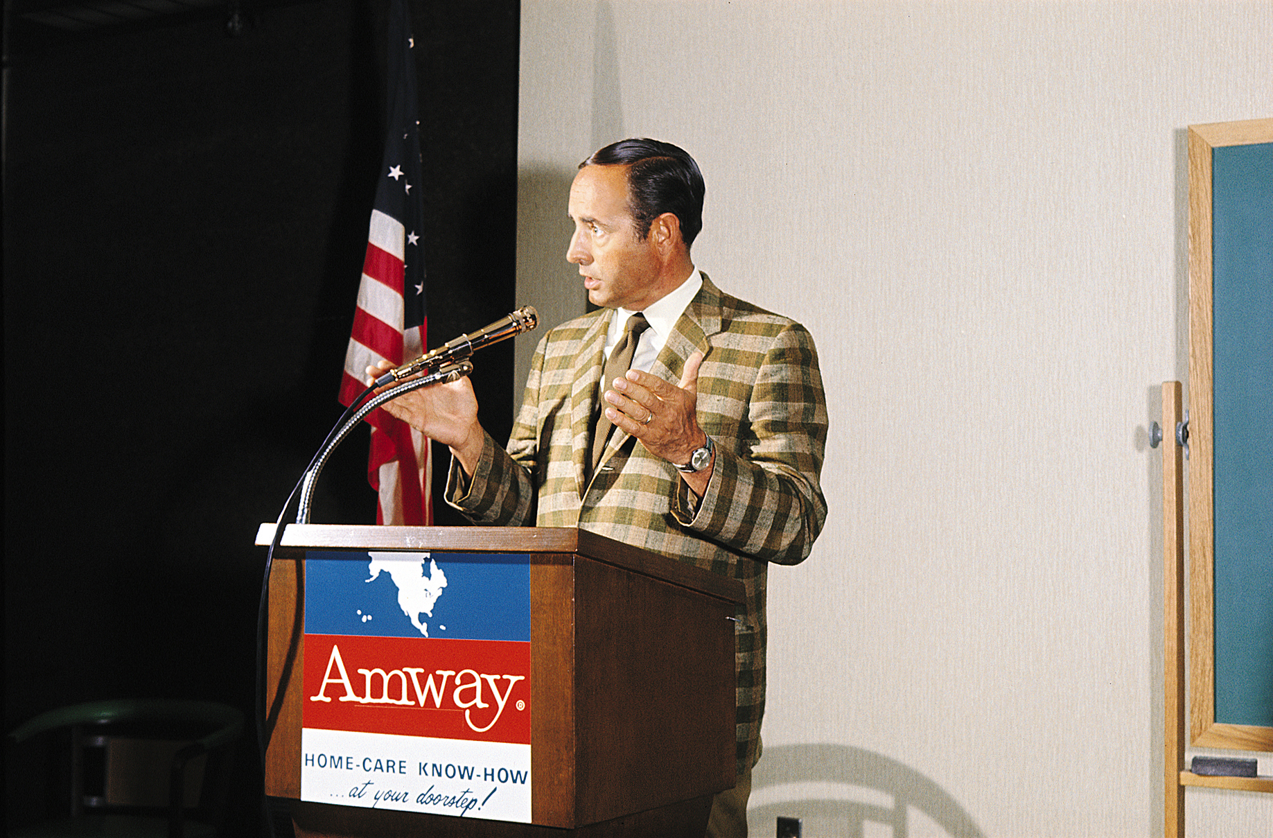 身着格纹西装的男士站在印有“Amway”标识的讲台后发表讲话，讲台标注标语“HOME-CARE KNOW-HOW ...at your doorstep!”，旁侧立有美国国旗。男士发言时手势生动、状态投入，现场氛围正式专业，为安利早期业务宣讲场景。