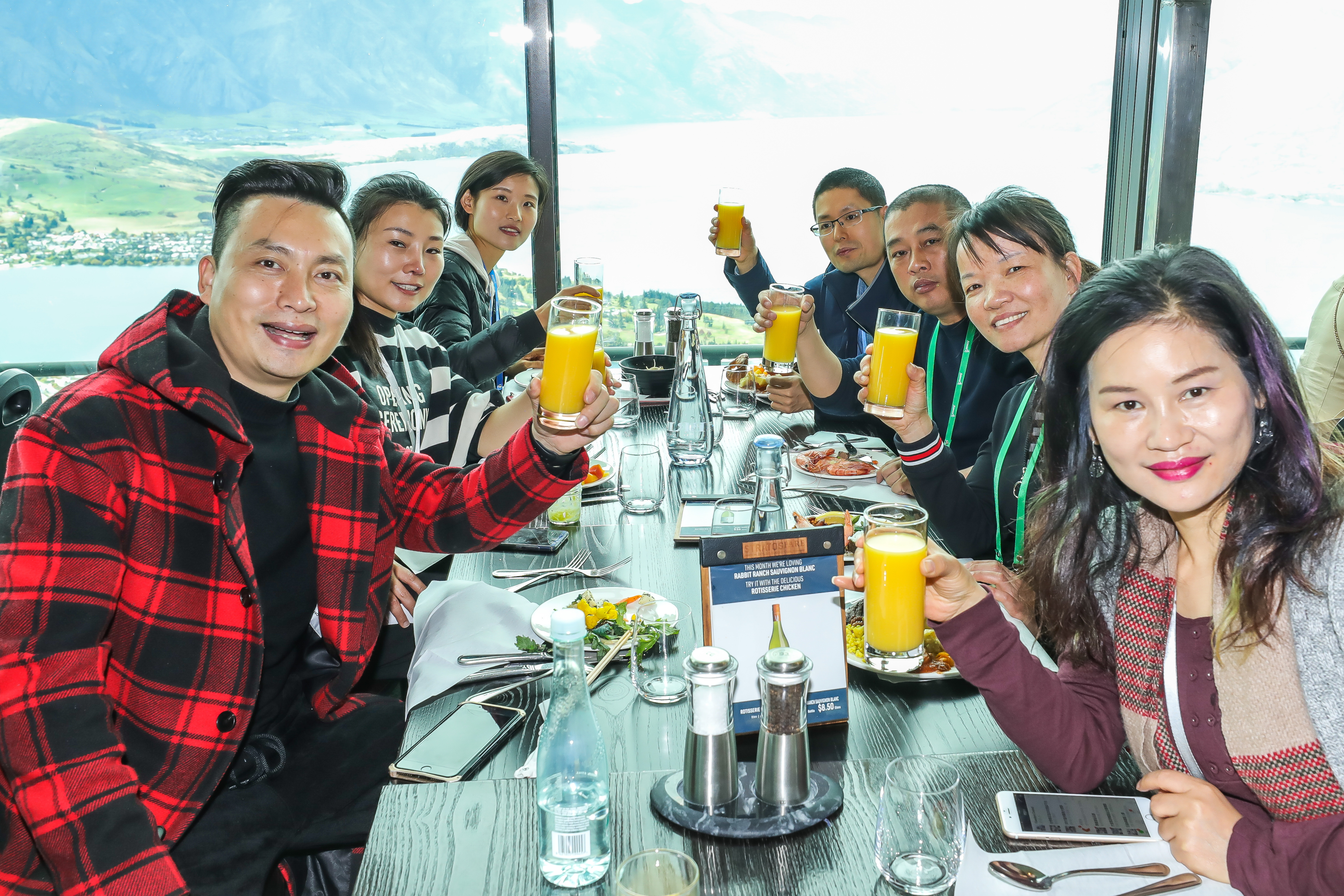 安利伙伴们围坐于临湖观景餐厅的餐桌旁，共同举着橙汁饮品看向镜头，脸上满是愉悦笑容。桌上摆放着餐食与餐具，窗外是开阔秀美的湖山风光，氛围轻松热烈，尽显伙伴们共聚的融洽与快乐。