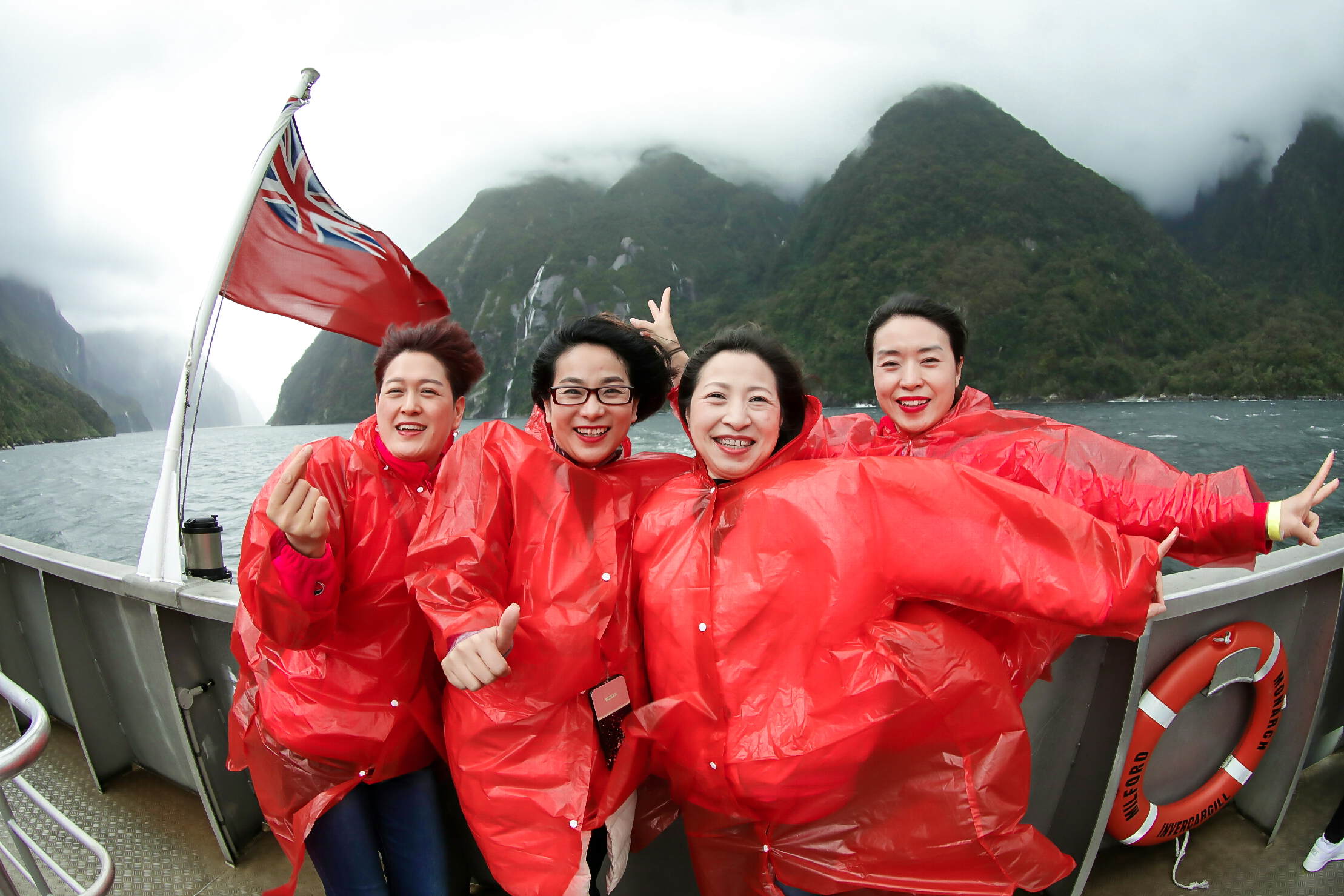 这是安利海外奖励之旅的游船游览环节，四位身着红色雨衣的安利伙伴站在游船甲板上，身后是云雾缭绕的峡湾青山与开阔水域，救生圈印有“MILFORD”“INVERCARGILL”“MONARCH”字样，大家笑容灿烂，姿态轻快，尽显旅途的愉悦与团队的蓬勃活力。