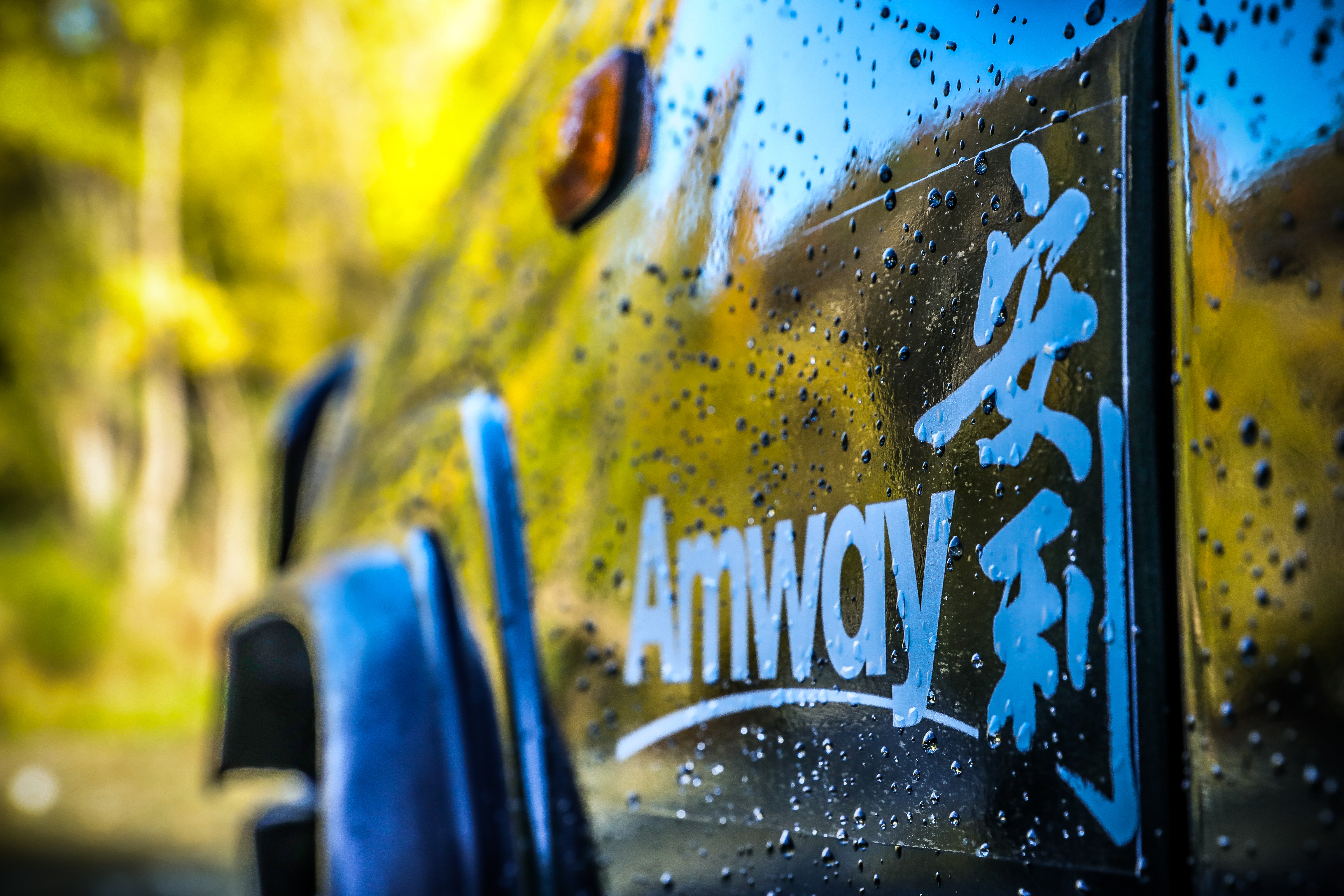 画面特写附着晶莹水珠的车身，其上清晰呈现“Amway”“安利”中英文品牌标识，背景是虚化的暖调户外自然景致，整体氛围清新明快，质感饱满，尽显安利品牌的鲜活质感与向上活力。