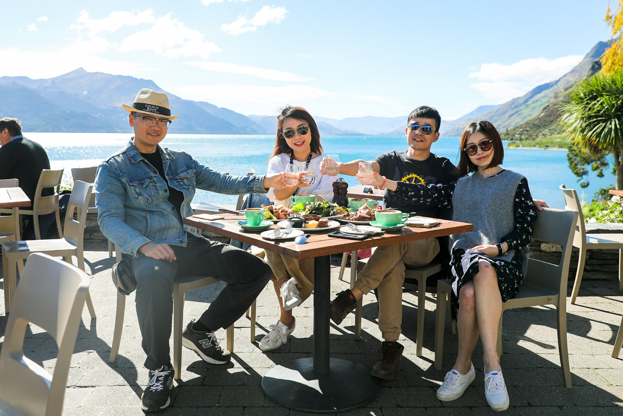 晴朗蓝天之下，澄澈湖泊与连绵远山相映成景，四位安利伙伴围坐湖景户外餐厅的餐桌旁，桌面摆放着餐食与饮品，众人笑容明朗一同举杯碰杯，氛围轻松愉悦，尽显伙伴同行的融洽惬意，传递积极向上的品牌温度。