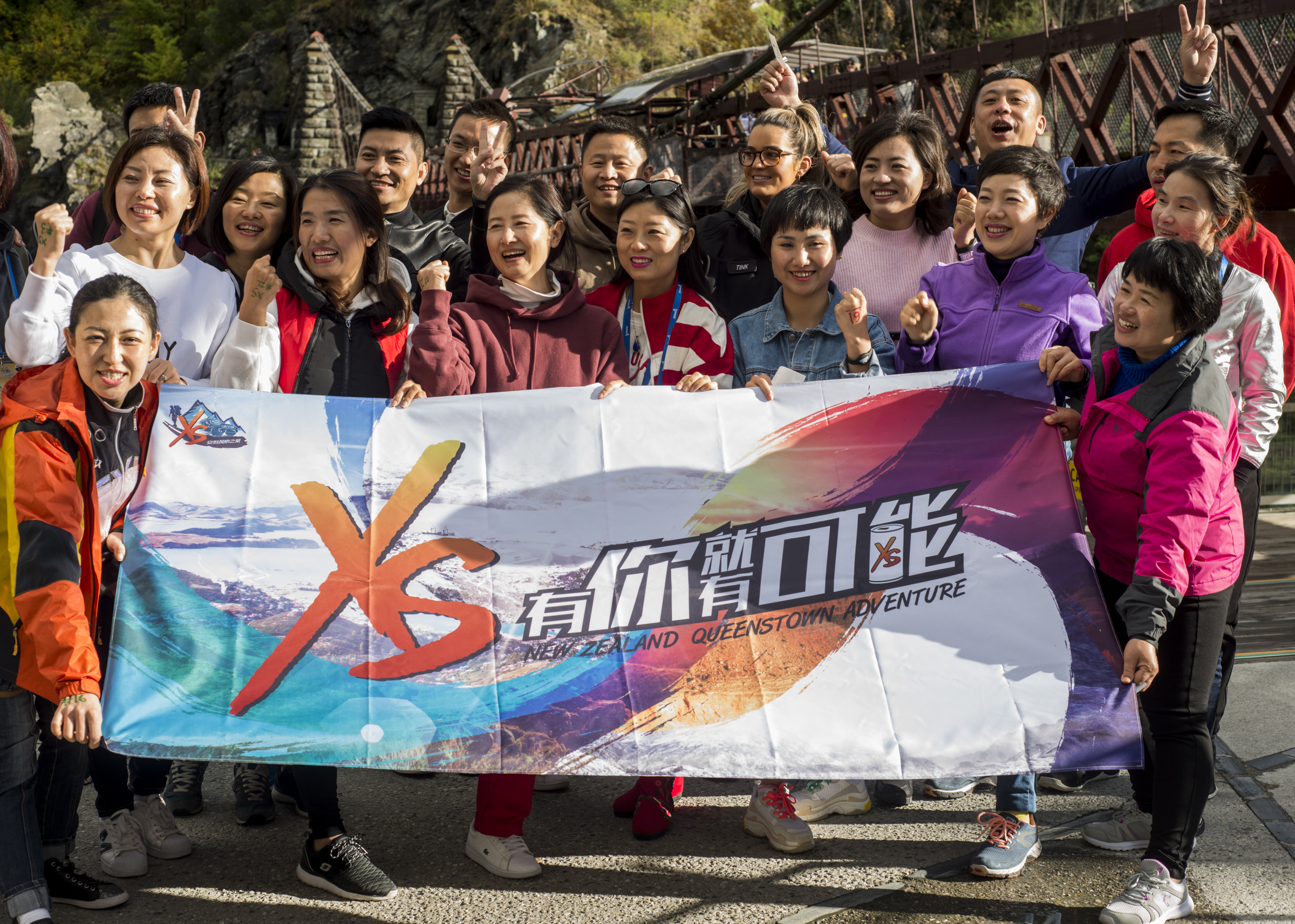 画面中一众安利伙伴齐聚新西兰皇后镇户外吊桥前，共同举着印有“XS 有你就有可能 NEW ZEALAND QUEENSTOWN ADVENTURE”字样的主题横幅，众人笑容明朗、姿态昂扬，氛围热烈活力十足，尽显团队凝聚力与安利积极向上的品牌风貌。