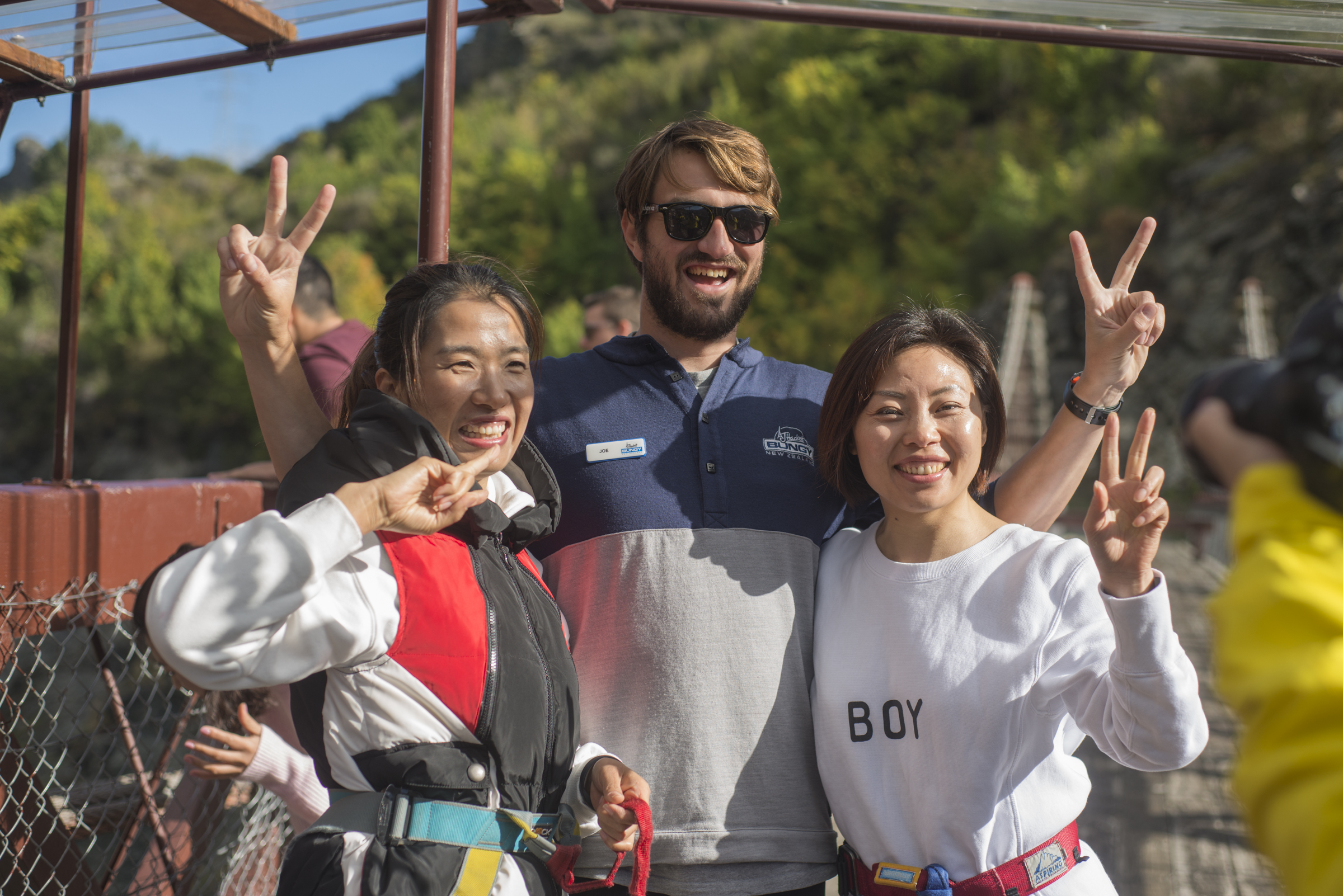 图片拍摄于安利海外研学行程的新西兰蹦极站点，两位穿戴好蹦极安全护具的安利伙伴，与身着印有“BUNGY NEW ZEALAND”工服、名牌标注“JOE”的站点工作人员亲密合影，三人纷纷比出剪刀手，笑容灿烂，氛围轻松明快，尽显安利伙伴勇于挑战、活力昂扬的精神风貌。