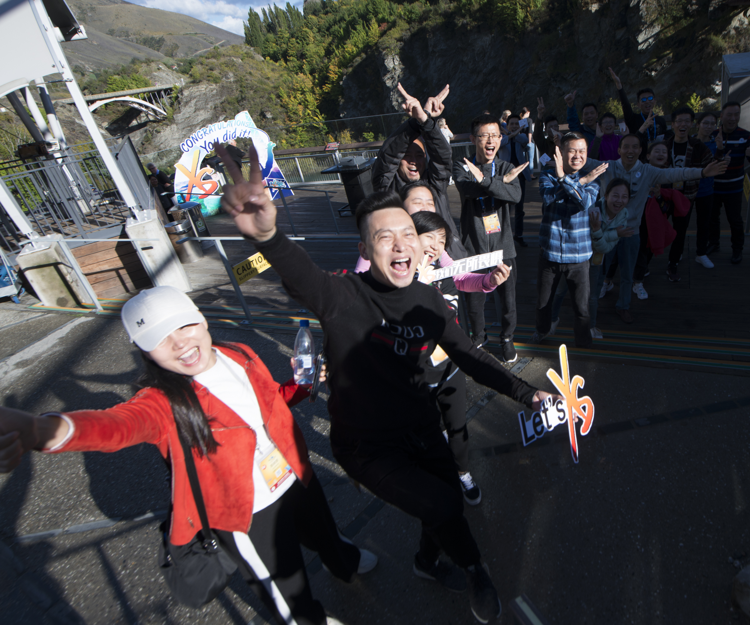 一众安利伙伴在户外山间平台集体合影，大家笑容灿烂、姿态昂扬，手持印有“Let's XS”“我们无所畏惧”的手牌，背景设有“CONGRATULATIONS You did it!”标识，现场氛围热烈欢快，尽显团队的蓬勃活力与昂扬斗志。