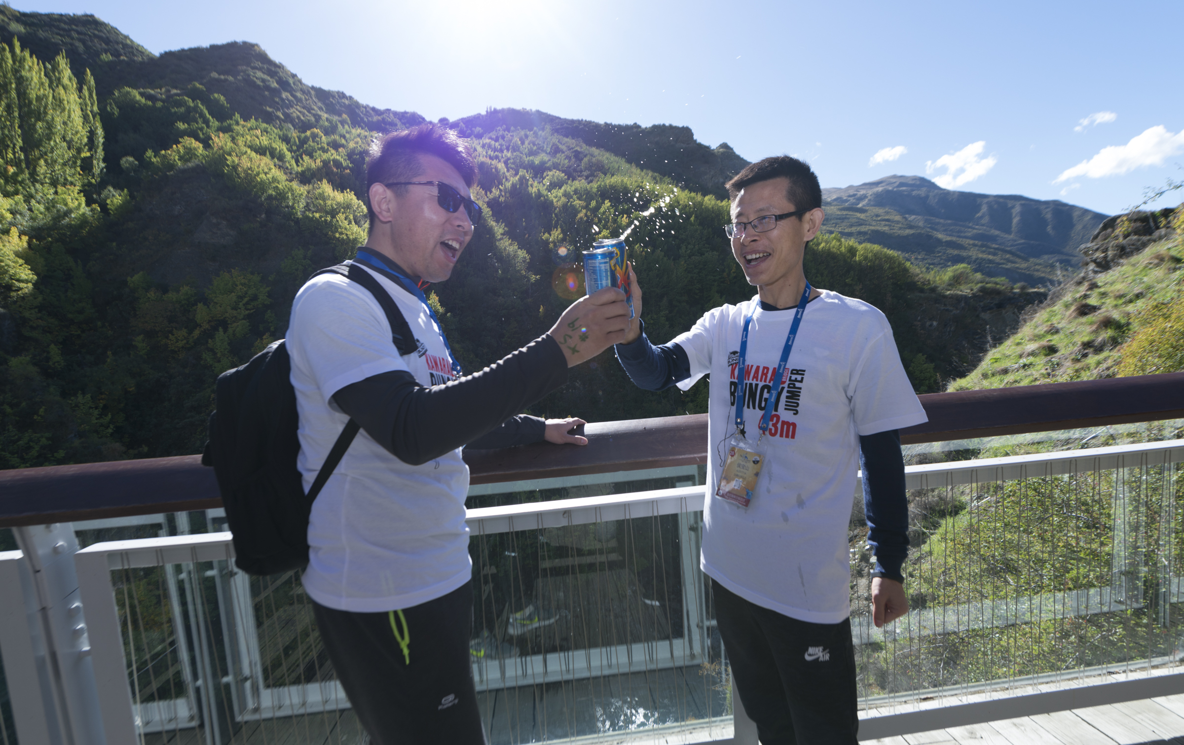 晴朗的山间观景台上，两位身着印有“KAWARA BUNGY JUMPER 43m”活动T恤的安利伙伴，完成蹦极挑战后开心举着饮料罐碰杯，饮品飞溅、二人笑容灿烂，氛围轻松热烈，尽显挑战自我的畅快与伙伴同庆的温暖，传递积极向上的风貌。