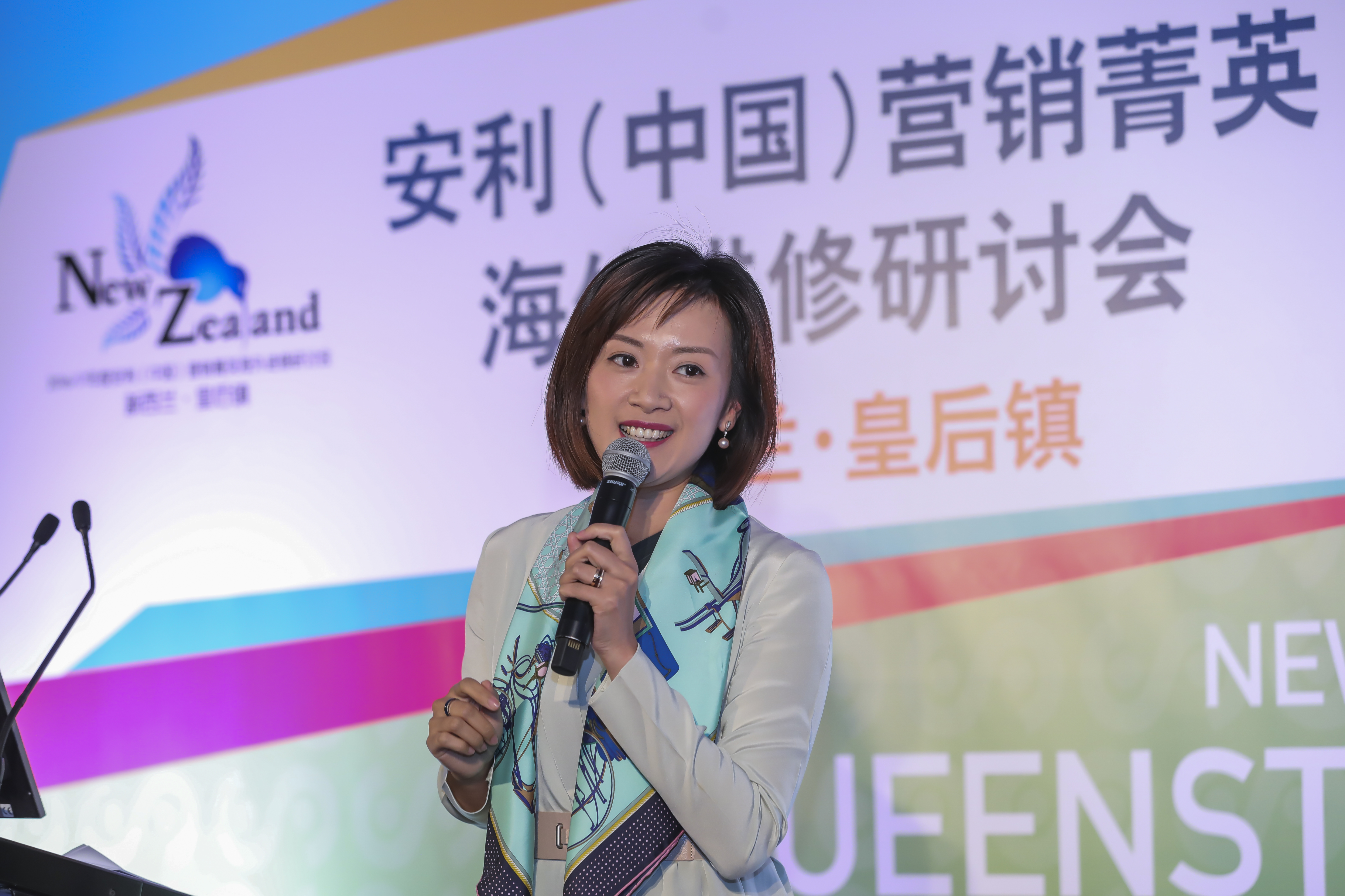 本场景为安利（中国）营销菁英海外研修研讨会新西兰皇后镇站现场，一位专业干练的女性发言人手持麦克风正在分享，她笑容亲和状态饱满，背景展板清晰呈现活动主题标识，现场氛围积极昂扬，尽显安利营销菁英的良好风貌。