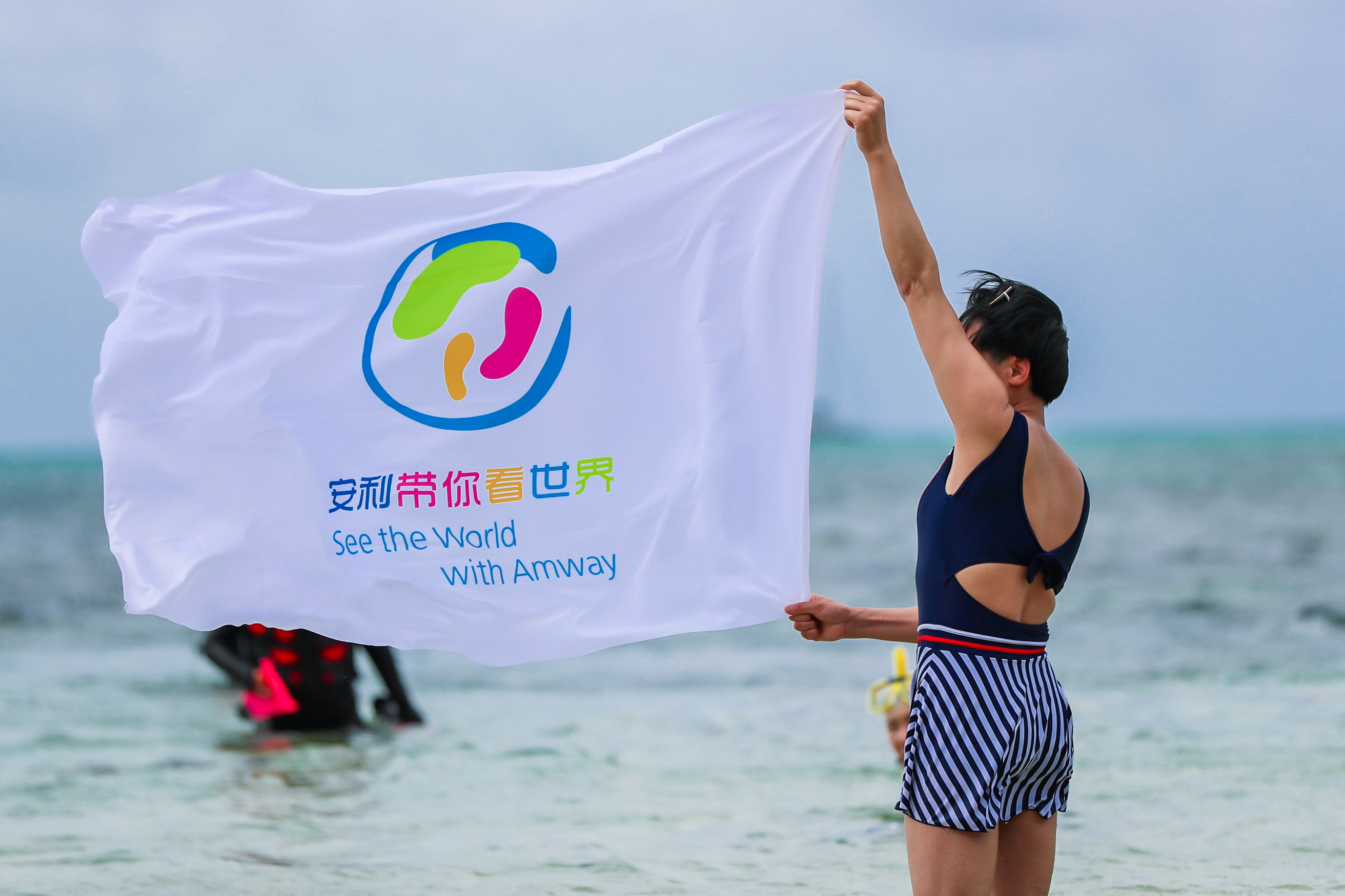 浅海区域中，身着泳装的安利伙伴高举印有“安利带你看世界”“See the World with Amway”字样的活动旗帜，旗帜在海风里舒展飘扬，背景海面还有其他参与活动的人员，整体氛围活力明快，尽显本次活动的轻松愉悦与安利伙伴的昂扬风貌。