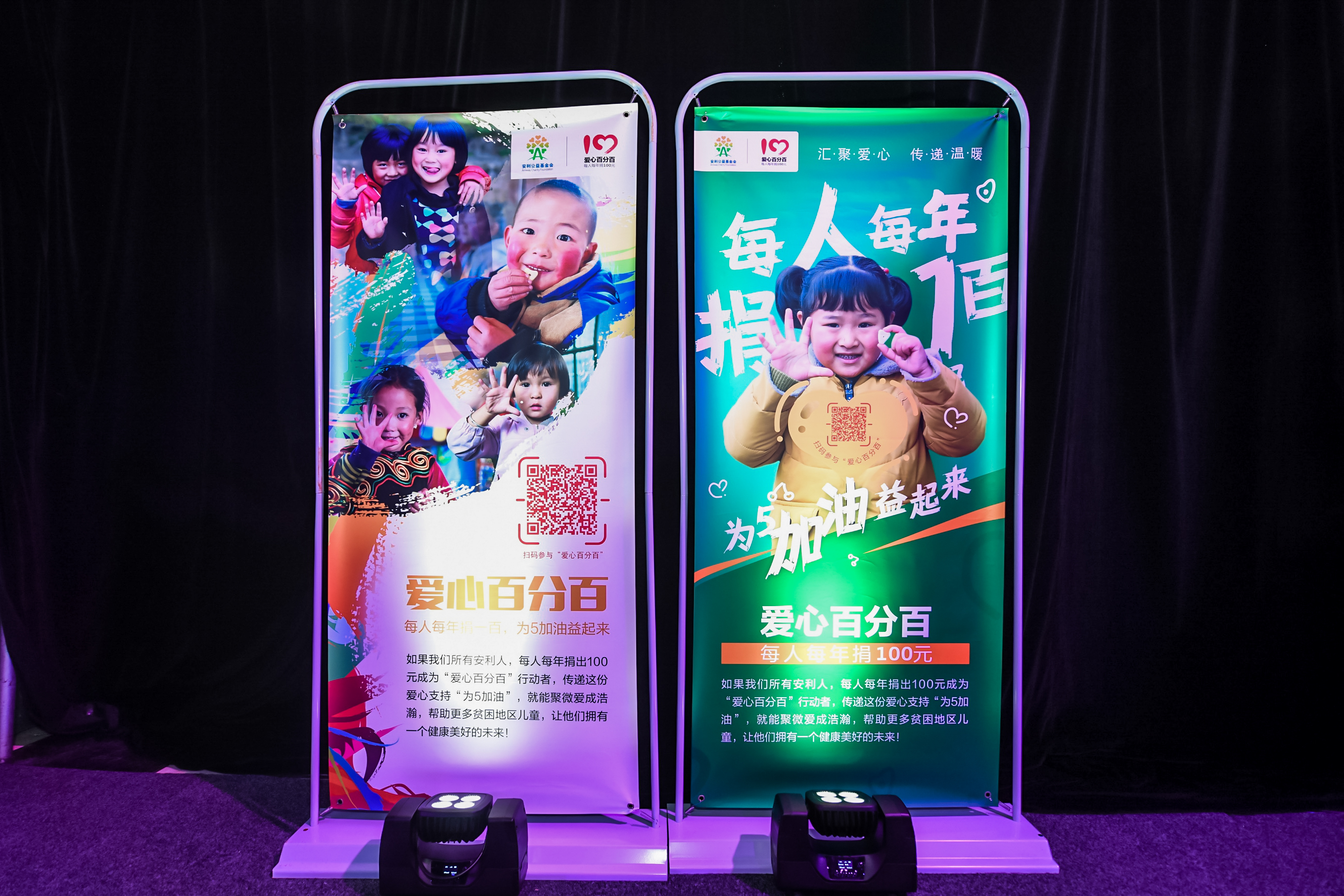 活动现场摆放两款安利“爱心百分百”公益宣传展架，印有“汇聚爱心 传递温暖”“每人每年捐100元”“为5加油益起来”等核心内容，搭配孩童笑脸视觉，倡议安利人参与捐款帮扶贫困地区儿童，传递公益向善的理念，氛围温暖正向。