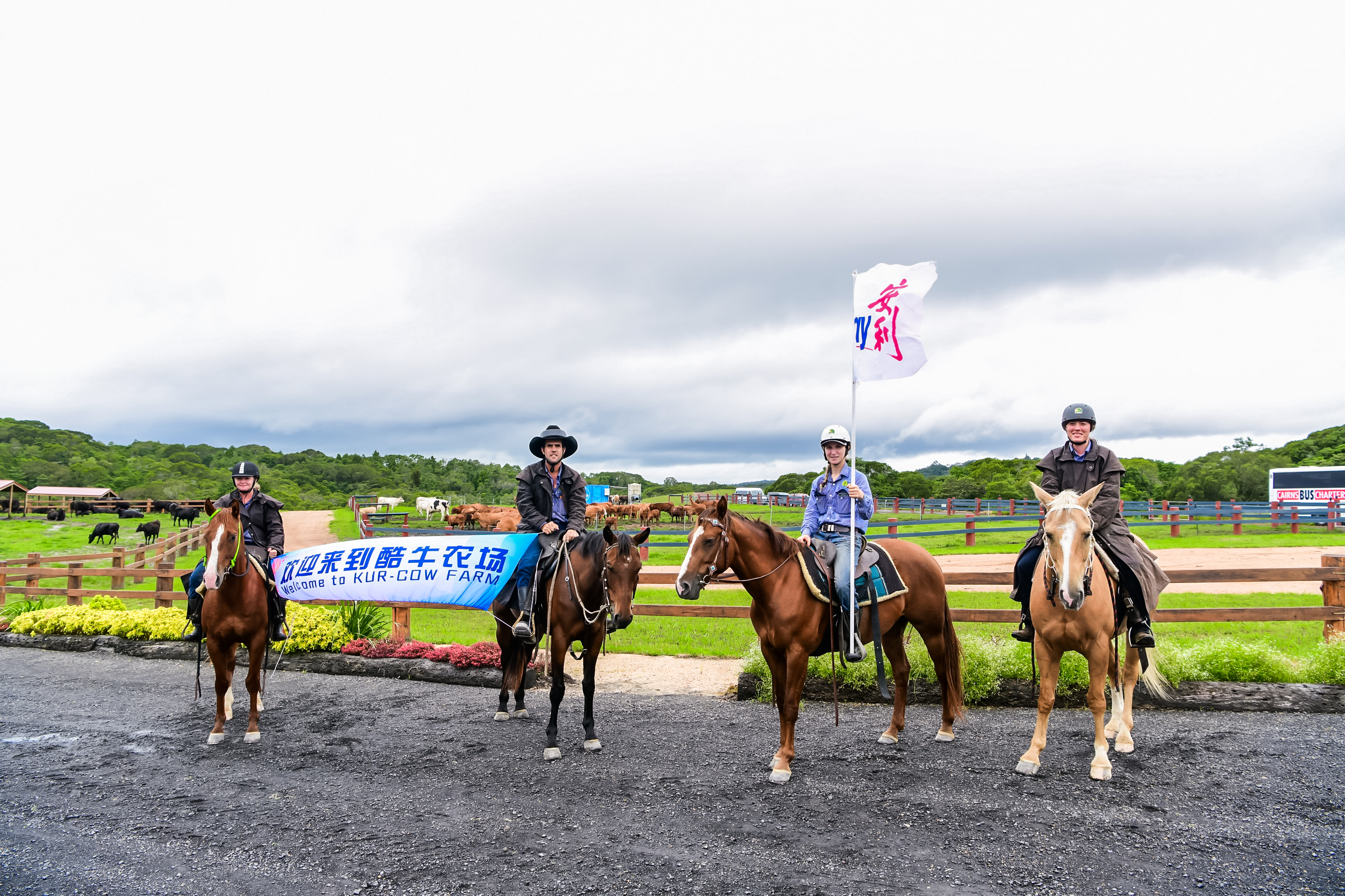 画面中四位骑手骑马列队，两人持“欢迎来到酷牛农场 Welcome to KUR-COW FARM”横幅，一人高举安利旗帜，背景是开阔青葱的酷牛农场牧场，氛围热忱友好，满含对安利团队到访的诚挚欢迎，尽显本次农场体验活动的活力基调。