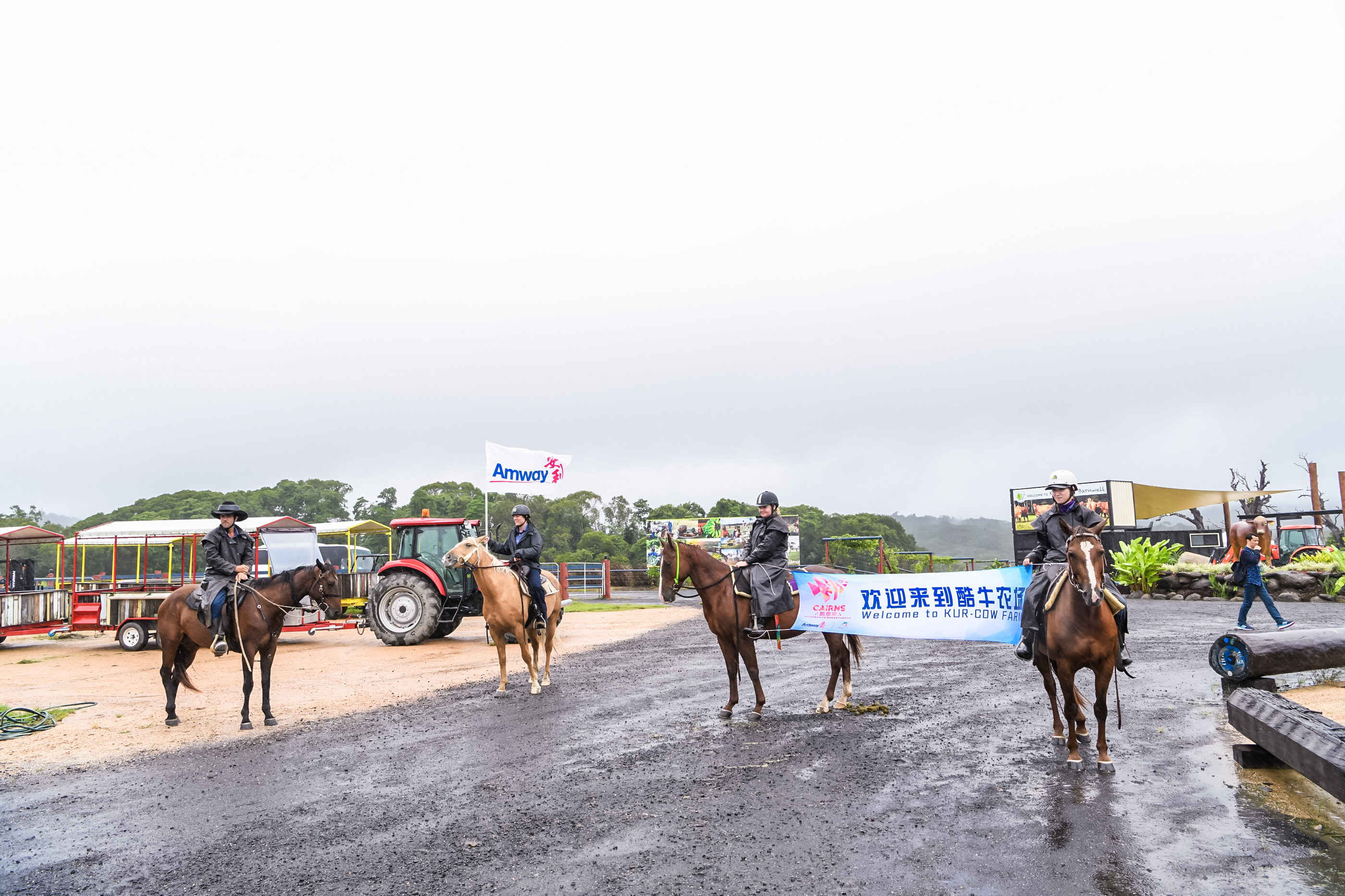 画面为安利酷牛农场体验活动迎宾现场，四位骑手骑马列队，一人手持印有“Amway”字样的品牌旗帜，两人拉着写有“欢迎来到酷牛农场 Welcome to KUR-COW FARM”的欢迎横幅，背景是开阔的农场场地与山林，仪式感满满，热情迎接参与活动的安利伙伴，氛围鲜活愉悦。