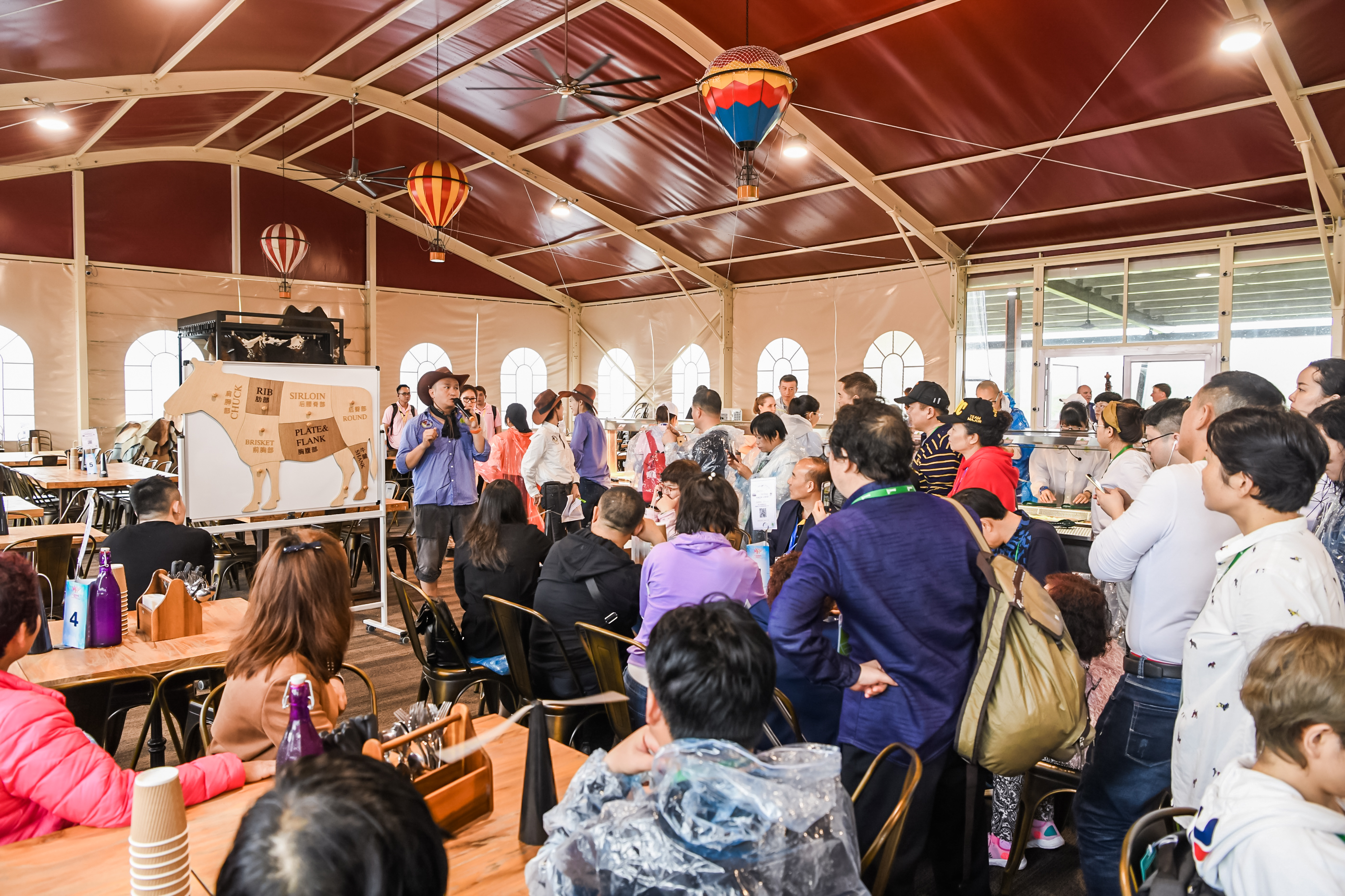 活动现场讲师正向参与活动的安利伙伴讲解牛肉部位知识，展示展板标注有SIRLOIN、RIB、CHUCK、BRISKET等各牛肉部位对应名称，在场人员专注聆听，氛围轻松热烈，是兼具趣味性与实用性的美食知识交流场景。