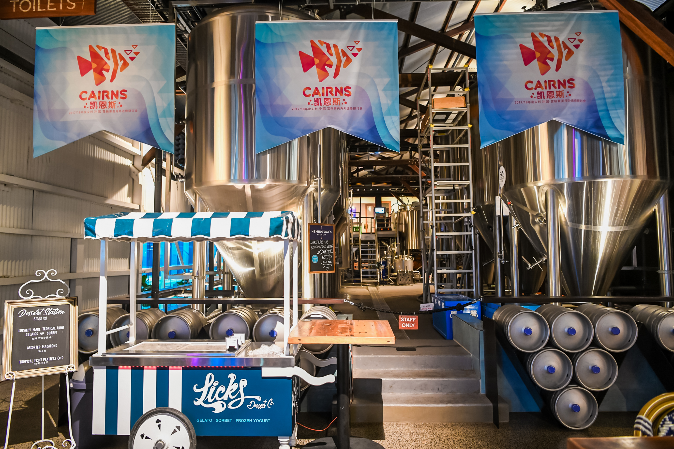 现场悬挂印有“CAIRNS 凯恩斯 2017/18年度安利(中国)营销菁英海外进修研讨会”的主题吊旗，场地为工业风精酿空间，排布着专业不锈钢酿造设备，一侧设置特色甜品站，氛围轻松舒适，为参会菁英打造了优质的休闲活动场景。