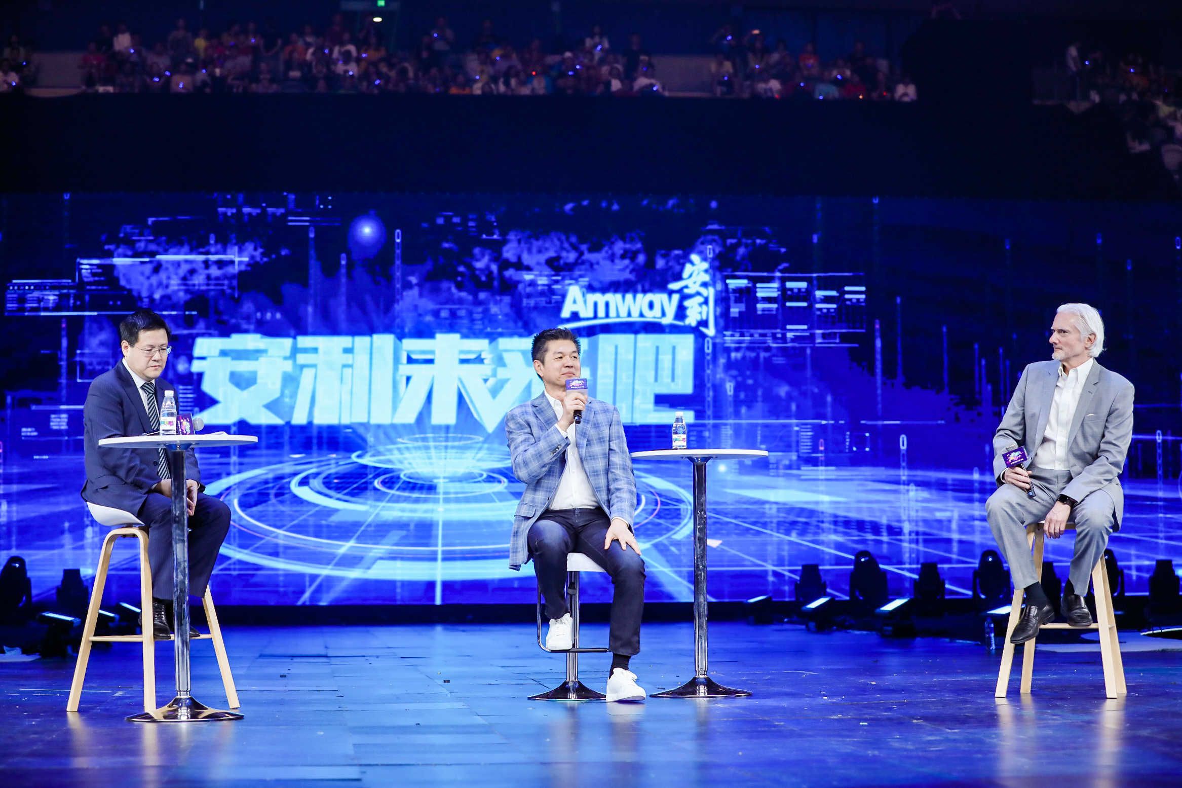 现场背景大屏印有“Amway 安利”“安利未来吧”核心字样，三位嘉宾坐于舞台高脚凳上参与主题研讨，中间嘉宾正持麦分享观点，舞台设计充满科技感，台下坐满参会观众，整体氛围专业热烈，展现出安利共探发展、畅想未来的积极风貌。