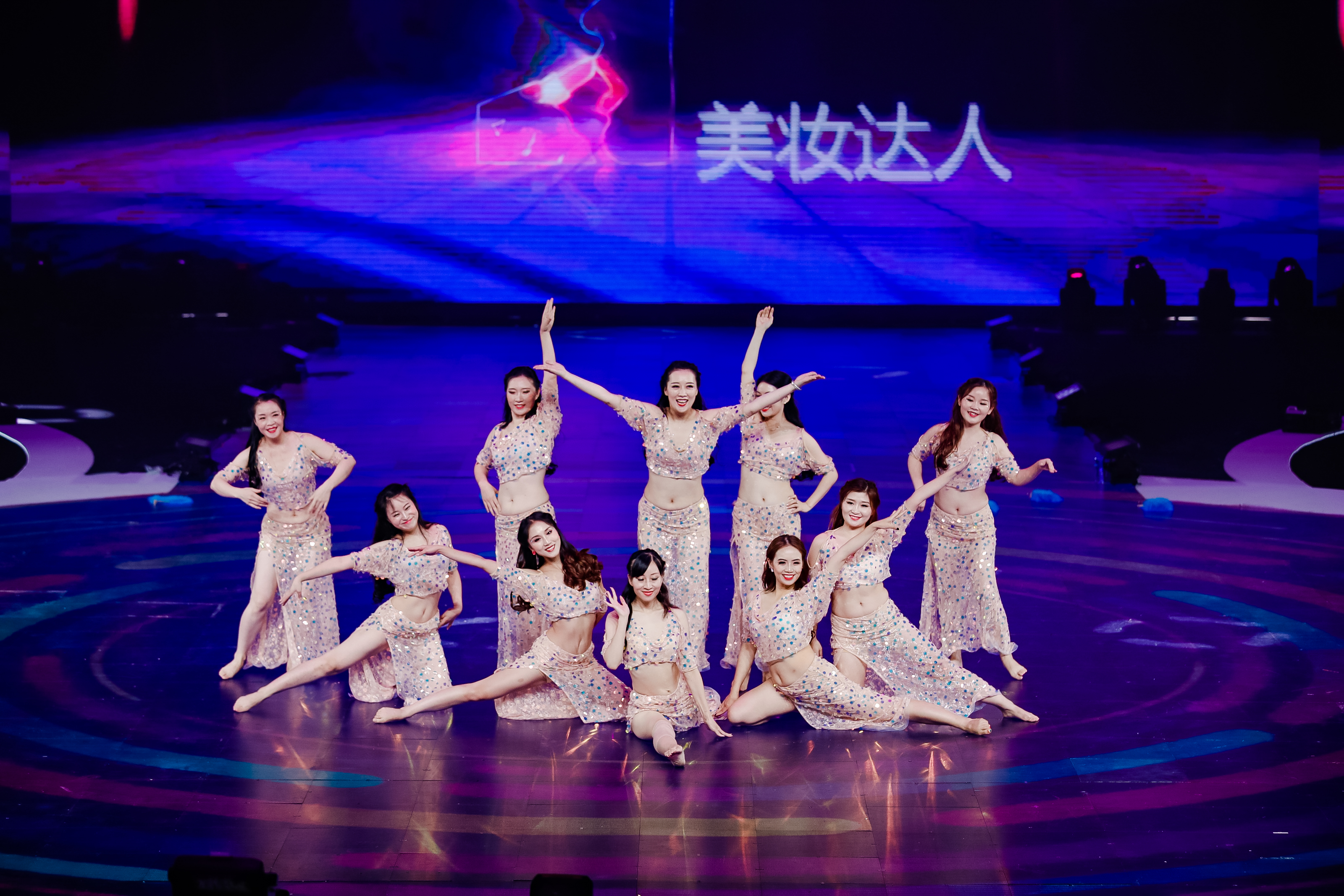 舞台背景大屏显示“美妆达人”字样，十余位身着缀彩亮片舞蹈服的安利伙伴，在紫调炫丽的舞台上摆出错落优美的造型，笑意明媚舒展，氛围活力热烈，充分展现出安利伙伴的多元风采与活动的鲜活氛围。