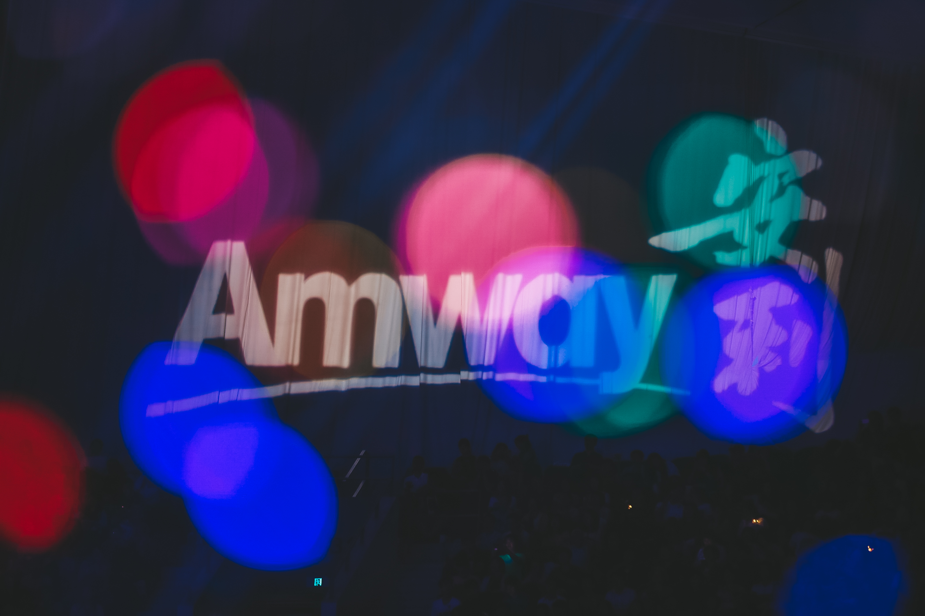 画面为安利活动现场场景，核心位置是醒目的Amway品牌标识，五彩梦幻的光斑点缀在画面中，台下坐满参会人员，现场灯光绚烂，氛围热烈庄重，尽显安利品牌活动的专业质感与活力。