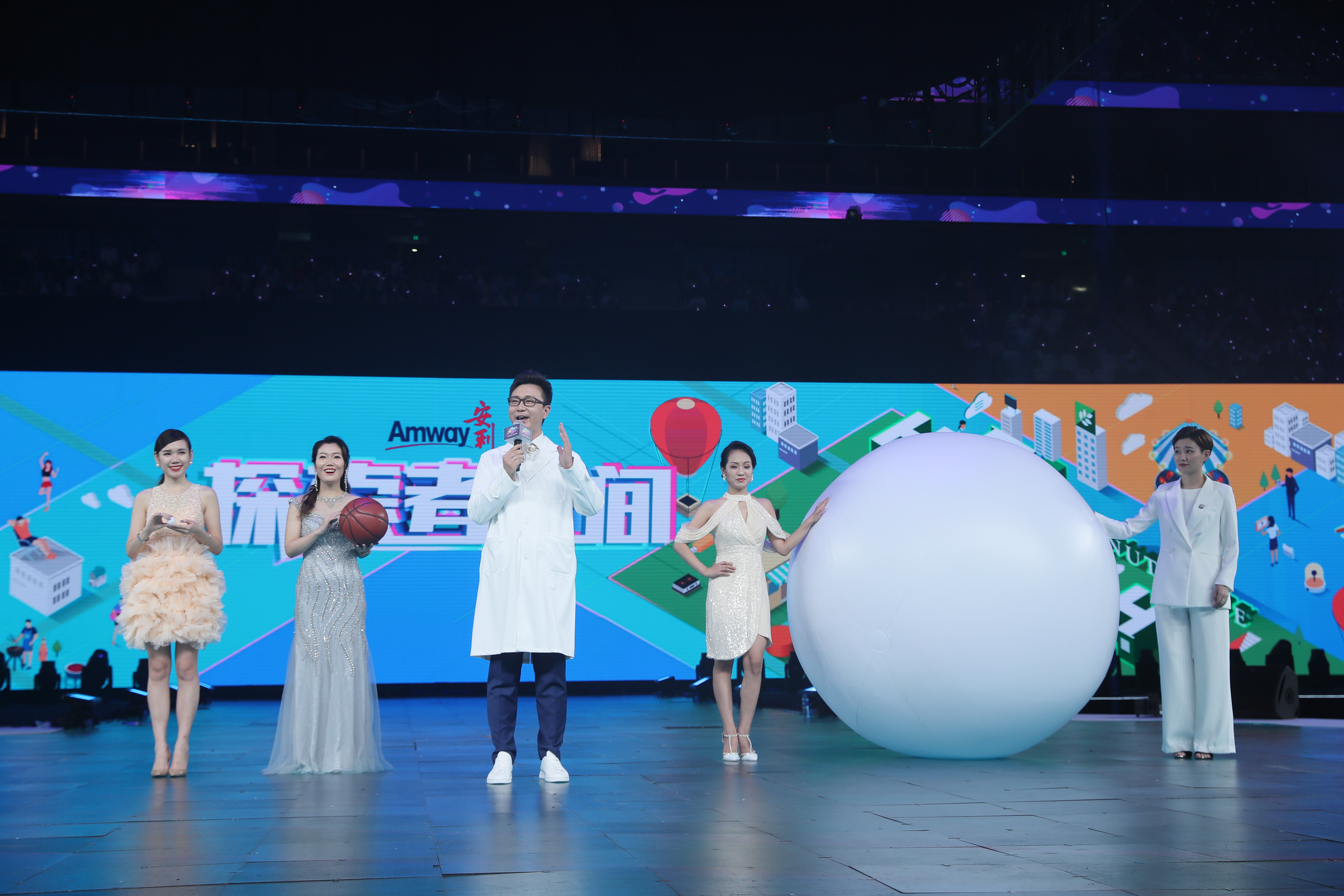 活动舞台背景印有“Amway安利”“探索者空间”字样，男主持人持麦正在发言，四位嘉宾分列舞台两侧，分别手持小物件、篮球，手扶巨型白色气球，现场氛围明快活跃，充分展现安利品牌活动的活力风貌，传递积极正向的品牌价值。