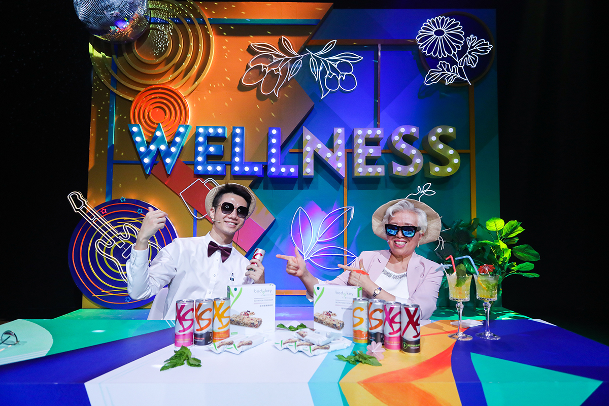 现场背景设置醒目“WELLNESS”灯牌，风格炫彩活泼。两位参与者佩戴草帽墨镜状态轻松愉悦，桌面陈列XS饮料、bodykey营养棒等安利健康产品，整体氛围活力欢快，充分传递安利健康乐活的品牌理念。